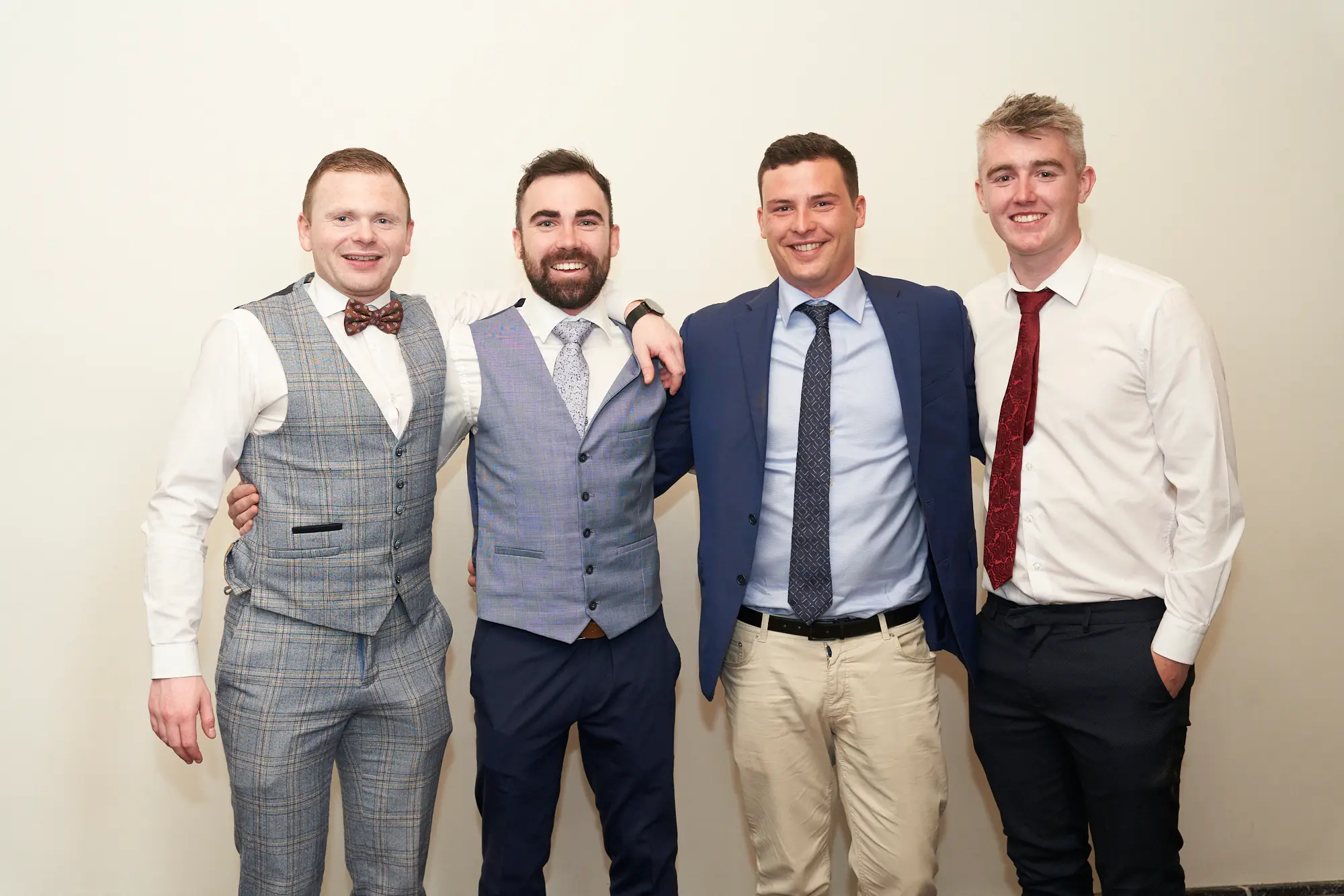 st kevins awards night pullman melbourne albert park four men group Four men pose together at the St. Kevin's Awards Night, standing side-by-side and smiling warmly at the camera. They are dressed in semi-formal evening wear including suits, waistcoats, and ties, with one man placing his arm around another's shoulder. The indoor function room features light beige walls, a dark carpet, and bright even lighting. Their relaxed, friendly poses and cheerful expressions convey a social and celebratory atmosphere, capturing a moment of camaraderie and enjoyment during the awards night event.