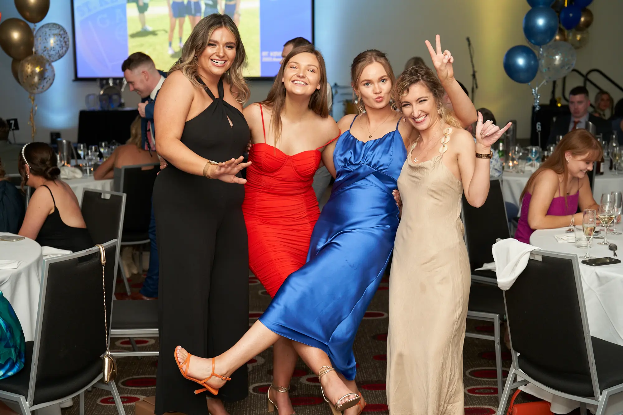 st kevins awards night pullman melbourne albert park four women group photo celebration Four women pose playfully for a group photo at St. Kevin's Awards Night at Pullman Melbourne Albert Park. From left to right: one wears a black jumpsuit with her hand on her hip, the next wears a red dress, the third wears a blue silk dress and kicks her leg up cheerfully, and the fourth wears a champagne-colored gown while flashing a double peace sign. Behind them, guests dine at round tables with white linens and black chairs, surrounded by blue and gold balloon decorations. The scene captures the joyful, fun, and celebratory energy of the awards night.