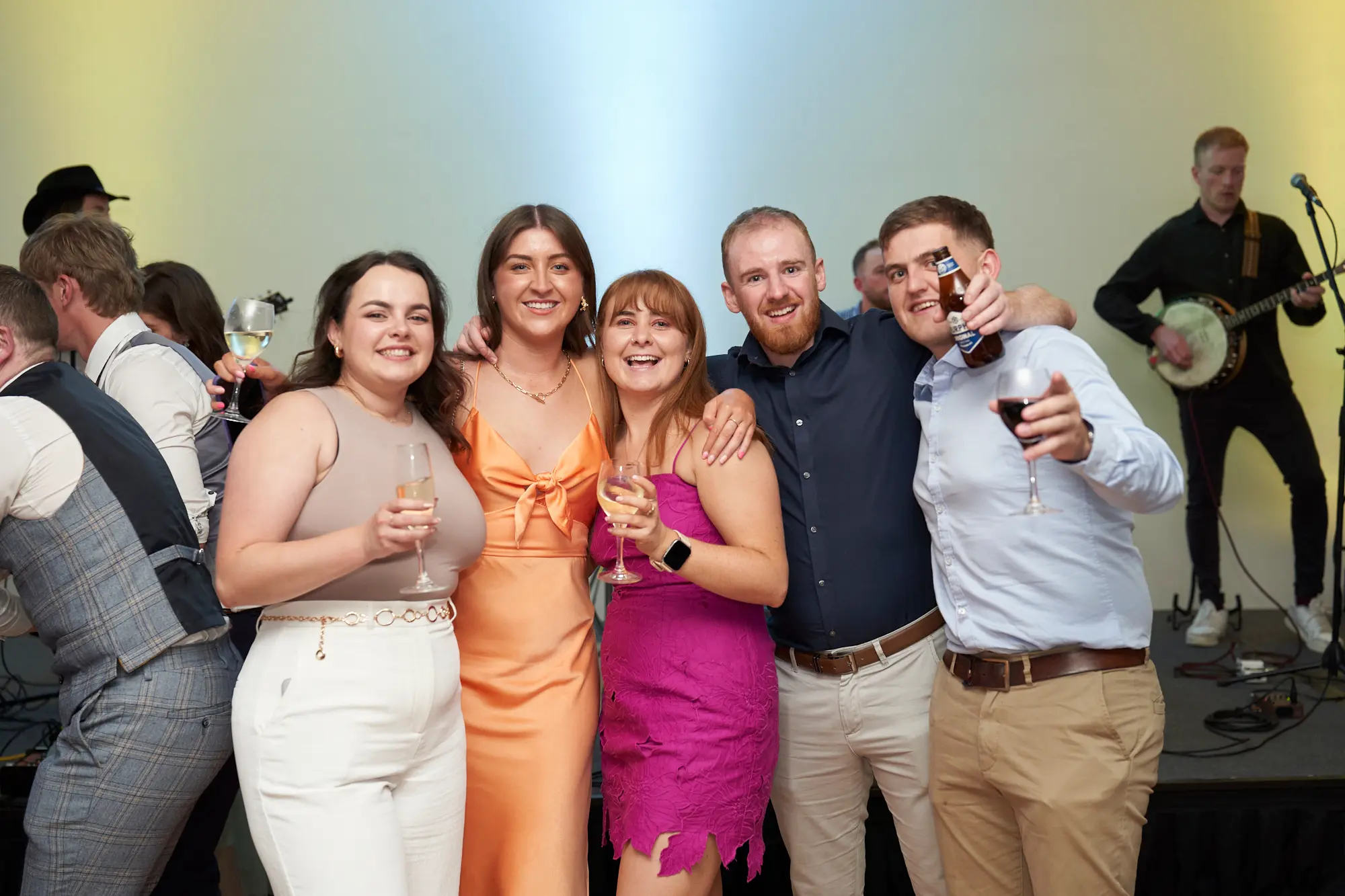 st kevins awards night pullman melbourne albert park friends celebration Six friends pose closely together at the St. Kevin's Awards Night, all smiling warmly at the camera. The group includes three women in vibrant dresses holding wine or champagne glasses, and three men dressed in smart-casual attire, one holding a beer bottle on his shoulder. Behind them, other guests mingle while a musician plays a banjo on stage. The dramatic yellow and green lighting, stage equipment, and dark flooring frame the lively, joyful, and social atmosphere, capturing genuine camaraderie and festive celebration during the awards night event.