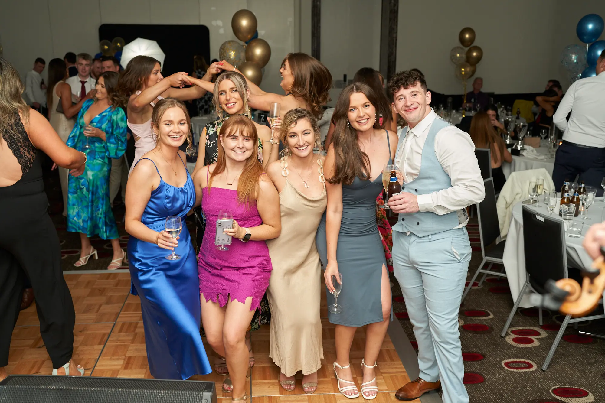 st kevins awards night pullman melbourne albert park friends dance floor Six friends pose closely together on the dance floor at the St. Kevin's Awards Night, all smiling warmly. The group includes women in royal blue, fuchsia, champagne, and grey dresses, and a man in a light blue three-piece suit holding a beer. Behind them, other guests mingle and dance, with one woman in a dark floral dress laughing and raising her arms. The wooden dance floor is surrounded by festive gold, blue, and confetti balloons, tables with glassware, and a violin on a stand. The scene captures joy, camaraderie, and a lively celebratory atmosphere.