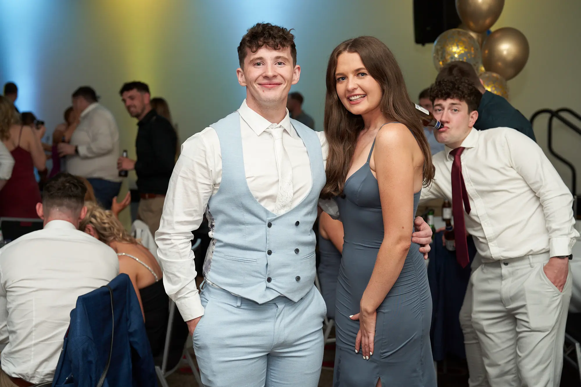 st kevins awards night pullman melbourne albert park friends posing during awards dinner Four young adults pose together for a social photo during St. Kevin’s Awards Night at Pullman Melbourne Albert Park. In the center, a man in a light blue three-piece suit smiles warmly beside a woman in a slate-grey satin slip dress with a side slit, her arm wrapped around his waist. To their right, another man in a white shirt and red tie playfully gestures toward the camera, while a fourth man in beige trousers stands casually nearby. Behind them, festive gold and blue balloons and warmly lit dinner tables create an inviting, celebratory atmosphere.