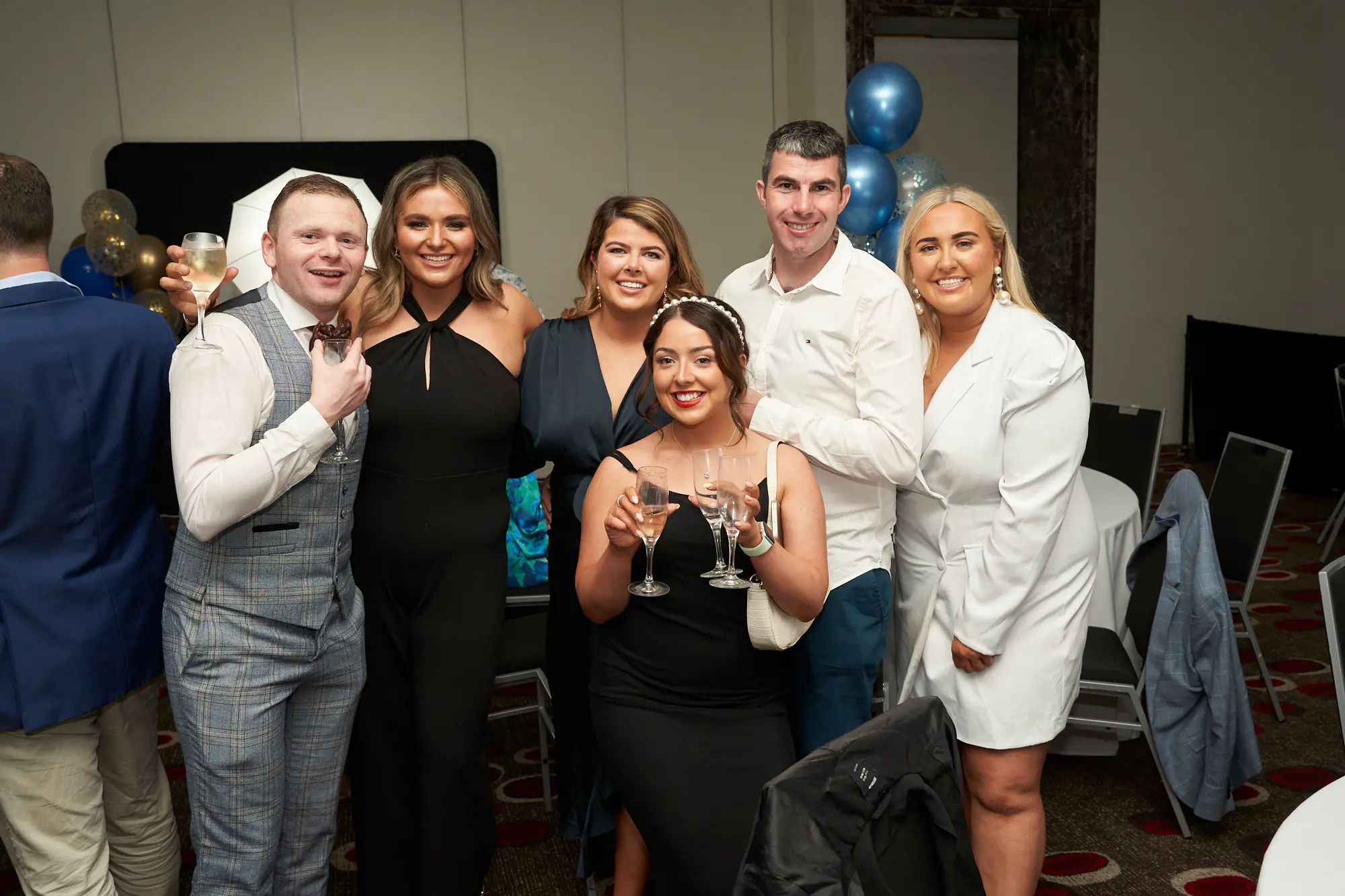 st kevins awards night pullman melbourne albert park group celebration ix well-dressed attendees pose closely together at the St. Kevin's Awards Night, all smiling brightly at the camera. Two individuals in the front row raise champagne flutes in celebration while the others smile warmly. The dark patterned carpet, clusters of gold and blue confetti balloons, and tables in the background frame the festive scene. Their joyful expressions, formal attire, and friendly poses convey camaraderie, celebration, and the lively, social atmosphere of an awards night gathering.