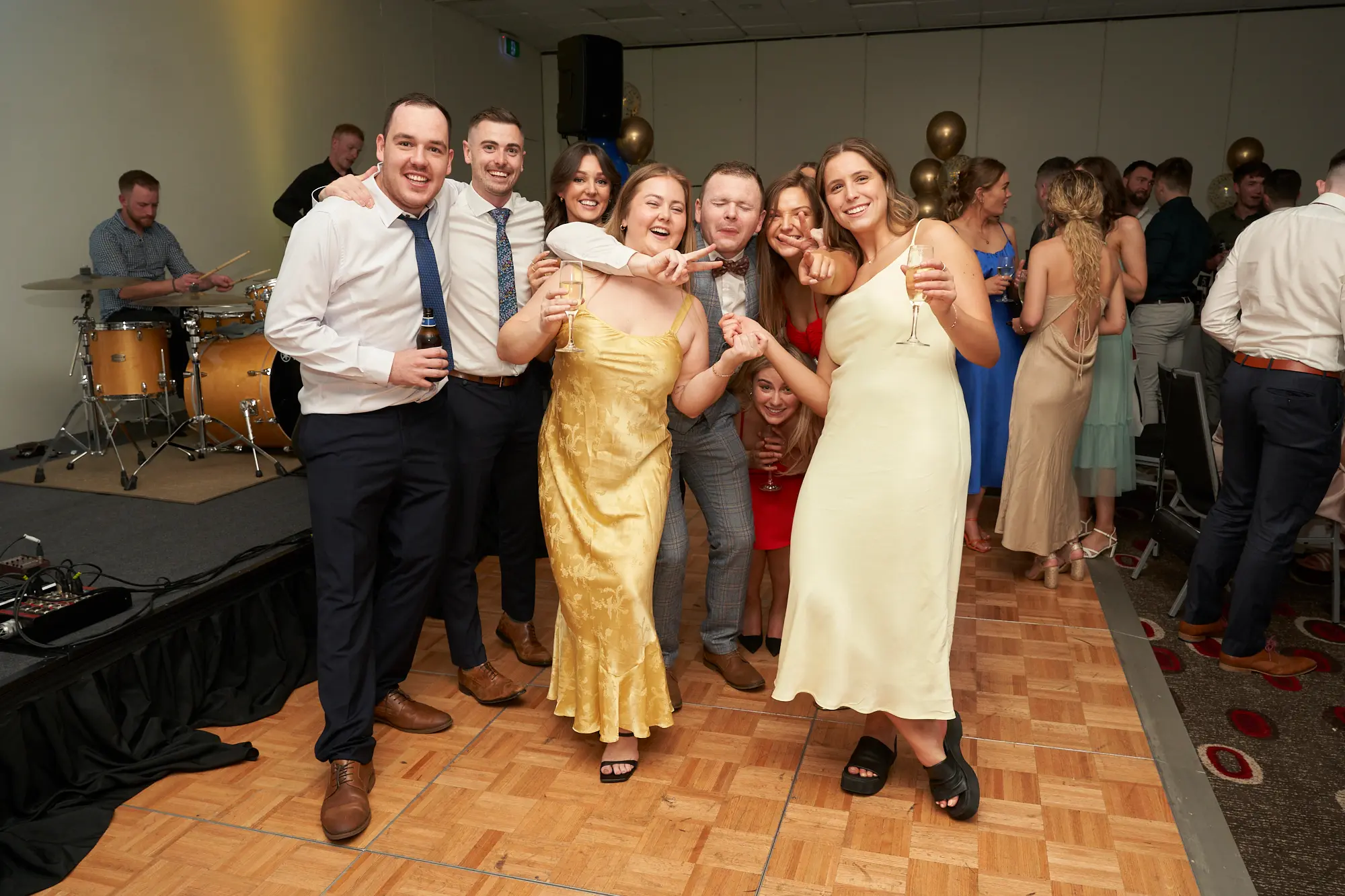 st kevins awards night pullman melbourne albert park group dance floor celebration A large, lively group of friends pose together on the dance floor during St. Kevin’s Awards Night at Pullman Melbourne Albert Park. The guests, dressed in elegant evening attire including gold, white, and navy outfits, smile brightly and hold drinks as they lean in for the photo. Behind them, other attendees dance, clap, and mingle under warm ambient lighting, while a drummer performs onstage near festive gold and blue balloons. The wooden parquet floor and laughter-filled expressions capture the vibrant, celebratory spirit of the post-awards party.
