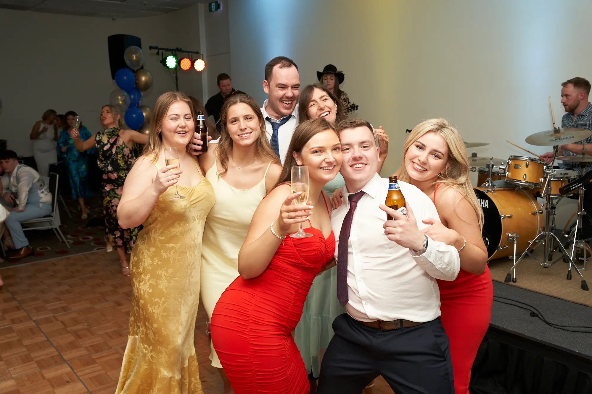 st kevins awards night pullman melbourne albert park group friends dance floor celebration A lively group of eight friends pose together on the dance floor during St. Kevin’s Awards Night at Pullman Melbourne Albert Park. Everyone is dressed in formal attire, smiling, laughing, and holding drinks, including beer bottles and champagne flutes. A woman in a gold dress and another in a red dress stand at the front, while others in white, mint green, and dark outfits join in the cheerful group pose. Behind them, a live band setup with a gold Yamaha drum set and festive gold and blue balloons creates a vibrant backdrop. The mood is joyful, energetic, and celebratory.
