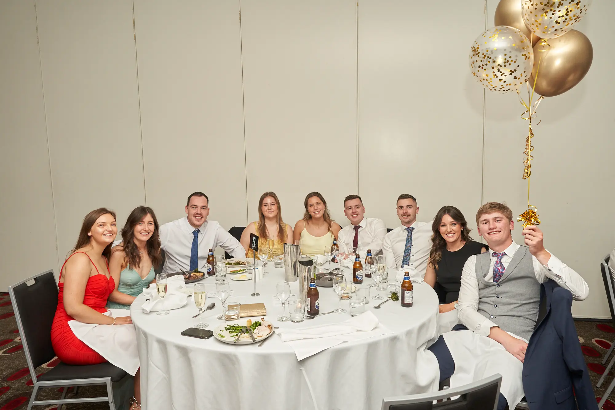 st kevins awards night pullman melbourne albert park group of eight dinner table 5 A group of eight young people pose for a photo at a dinner table during St. Kevin’s Awards Night at Pullman Melbourne Albert Park. The man on the far right, wearing a grey vest, holds a cluster of gold balloons, while the others smile at the camera. Plates, glasses, and bottles of beer and water are set on the table, which is marked with table number “5.” The background shows a well-lit banquet hall with light-colored walls and a red patterned carpet. The scene captures the joyful, social, and celebratory atmosphere of the evening.