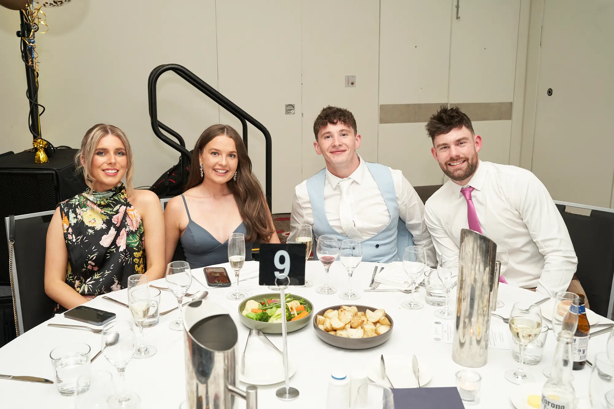 st kevins awards night pullman melbourne albert park group of four dinner table 9 Four people smile for the camera while seated together at a dinner table during St. Kevin’s Awards Night at Pullman Melbourne Albert Park. From left to right: a woman in a black floral dress, a woman in a blue-grey dress, a man in a light blue vest over a white shirt, and a man in a white shirt with a bright pink tie. The table, marked with number “9,” is set with plates of food, glasses, a small silver decoration, and a bottle of Corona beer. The scene captures the festive, social, and celebratory atmosphere of the evening banquet.