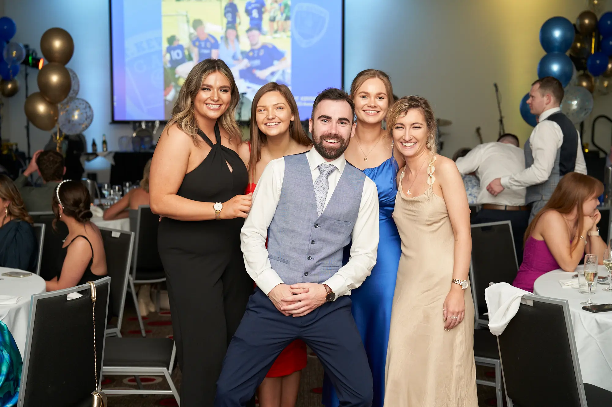 st kevins awards night pullman melbourne albert park group photo five guests celebration A lively group of five guests pose for a photo at St. Kevin's Awards Night at Pullman Melbourne Albert Park. A man in the center crouches slightly, smiling broadly in a grey vest, white shirt, and tie, surrounded by four women dressed in elegant gowns—one in black, one in red, one in blue, and one in a champagne-colored halter dress. Behind them, the banquet hall is filled with guests seated at round tables draped in white linens, accented by blue and gold balloons and a large projector screen displaying a team image. The scene radiates warmth, camaraderie, and celebration.