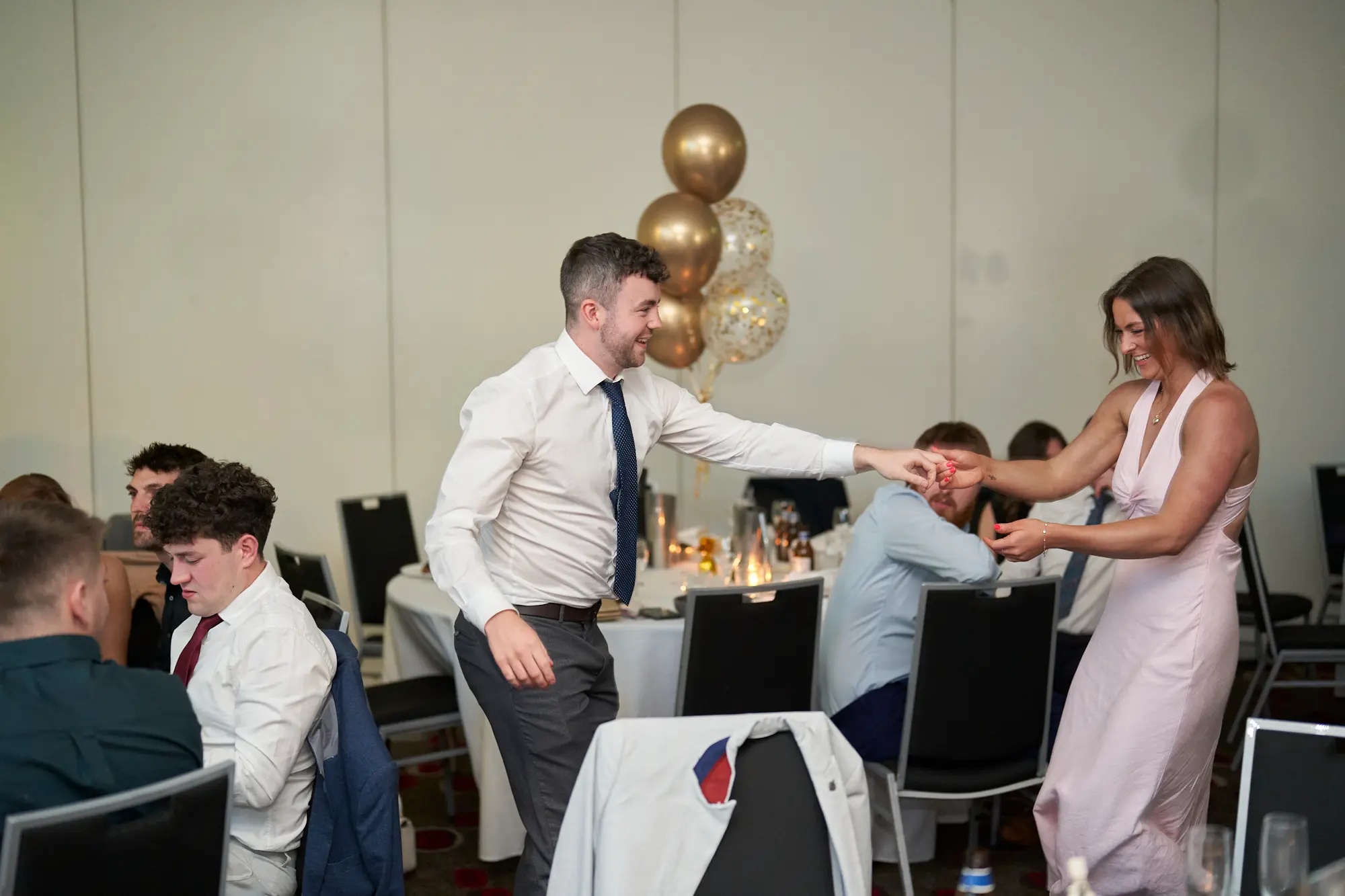 st kevins awards night pullman melbourne albert park guest interaction During St. Kevin's Awards Night, a man in a white shirt and navy polka-dot tie reaches out to a woman in a pink satin slip dress, smiling warmly as they engage near a table. Surrounding them, formally dressed guests sit at white-clothed tables, some holding drinks, while festive gold and clear confetti balloons add celebratory flair. The dark patterned carpet, neutral walls, and scattered chairs create a lively, social backdrop. This candid moment captures joyful interaction, signaling the transition from dinner to dancing and highlighting the warm, energetic atmosphere of the awards night celebration.