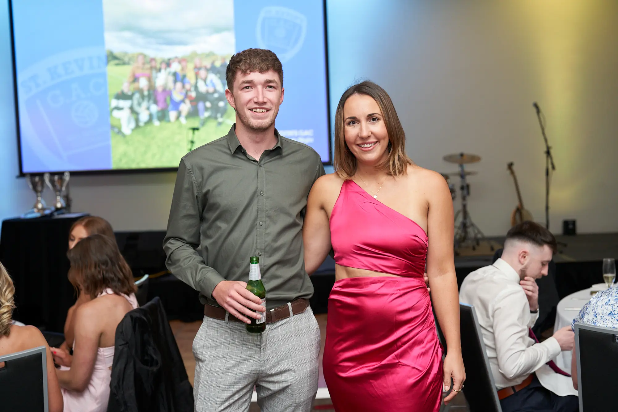st kevins awards night pullman melbourne albert park guest portrait beer trophy A man and a woman pose together during the annual St. Kevin's Awards Night, smiling brightly at the camera. The man wears a light olive-green button-up shirt and grey checkered trousers, holding a green glass beer bottle, while the woman wears a vibrant pink one-shoulder dress with a side cutout. Behind them, a table displays silver trophies and speakers, and a large projection screen shows a sports team with the text “ST. KEVIN'S G.A.C.”. A gold drum set and string instrument are visible to the right. The setting conveys a lively, celebratory awards night atmosphere.