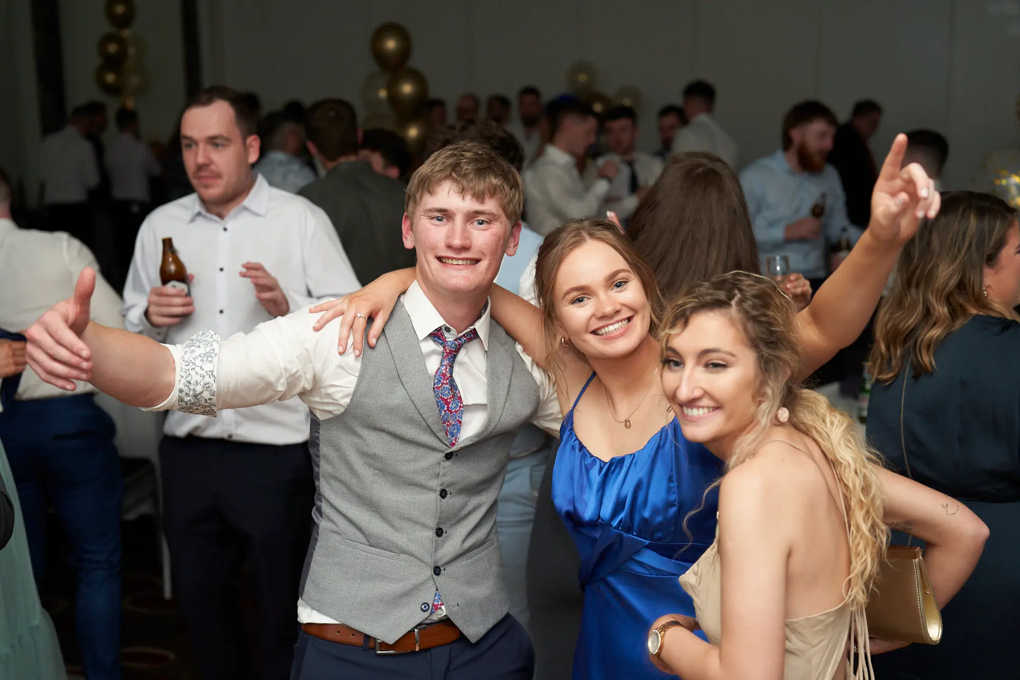 st kevins awards night pullman melbourne albert park guests celebrating together on dancefloor A lively group of young adults pose together during St. Kevin’s Awards Night at Pullman Melbourne Albert Park. In the center, a man in a grey waistcoat and patterned tie wraps his arms around two women — one in a blue dress raising her arm in celebration and another in a light gold dress smiling warmly. Beside them, a man in a white shirt gives a cheerful thumbs-up while holding a beer. Behind the group, elegantly dressed guests mingle beneath gold and blue balloons. The image radiates joy, camaraderie, and the festive spirit of the evening’s celebrations.