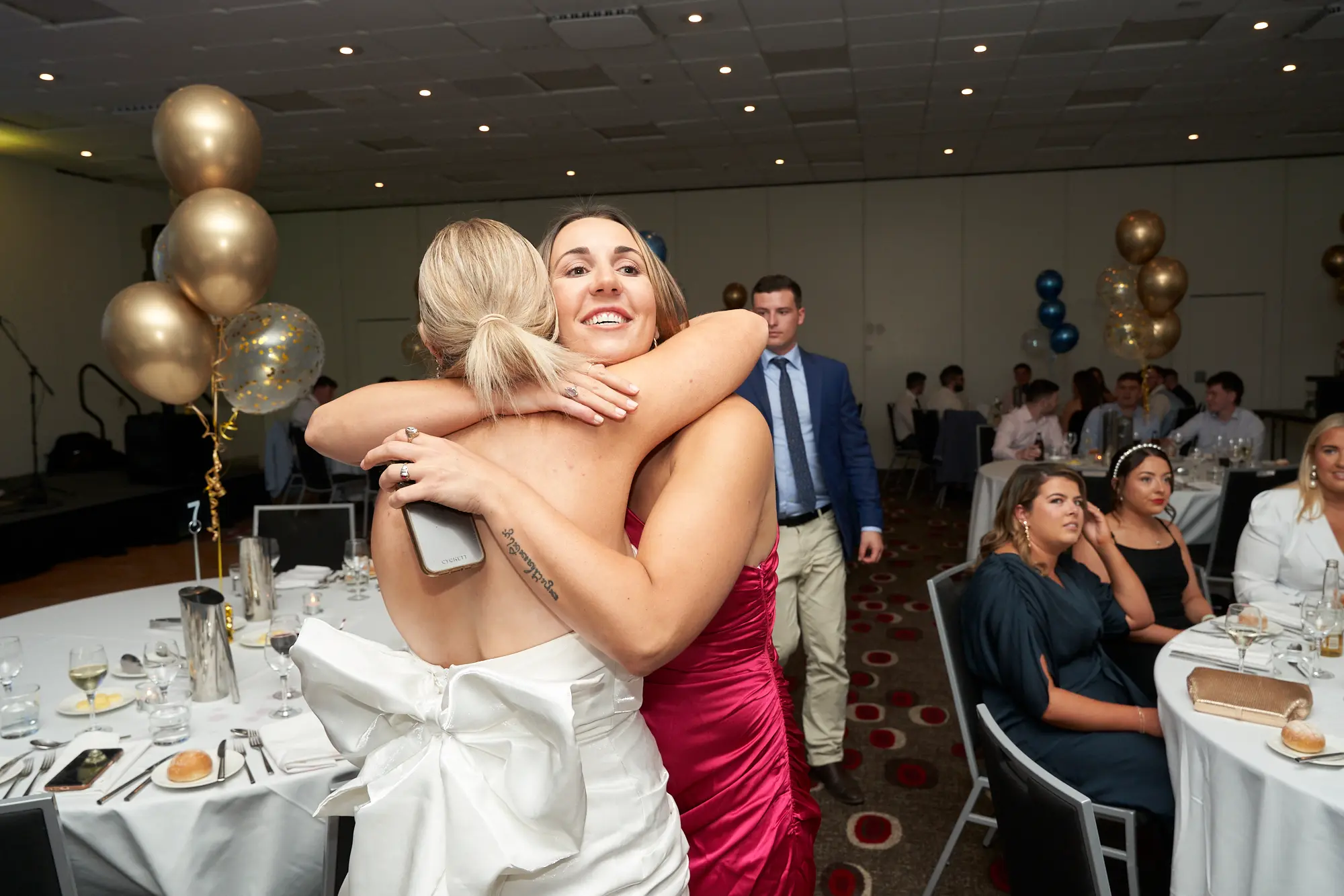 st kevins awards night pullman melbourne albert park guests embrace celebration Two women share a warm embrace during St. Kevin's Awards Night at Pullman Melbourne Albert Park. The woman on the left, seen from behind, wears a white strapless dress with a large bow, while the woman on the right, smiling toward the camera, wears a magenta gown. Behind them, guests sit at round dinner tables covered in white linens, surrounded by gold and clear balloons. A man in a suit stands near the center, and a stage setup is visible to the left. The scene captures the joyful, elegant, and heartfelt atmosphere of the awards night celebration.