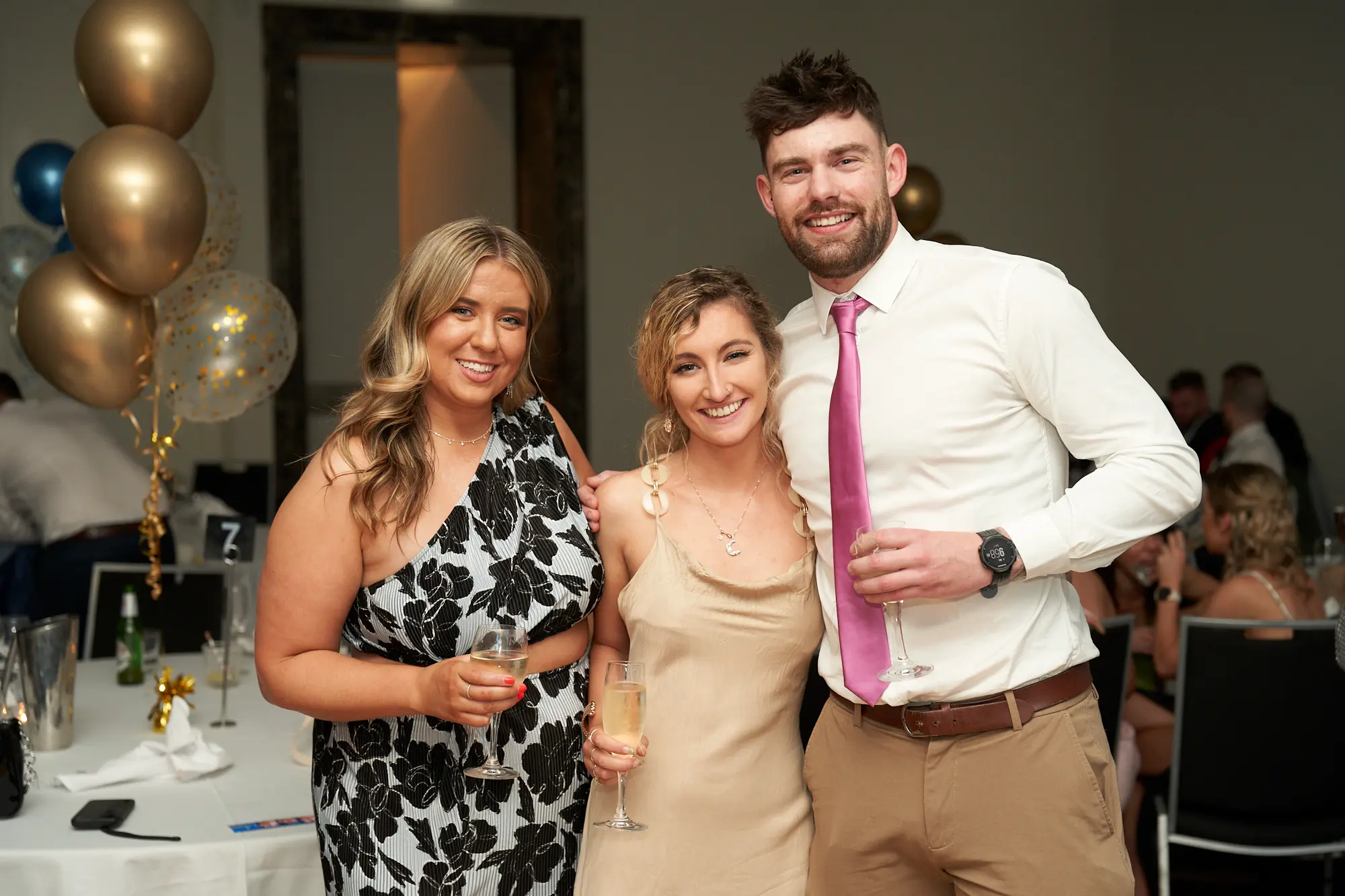 st kevins awards night pullman melbourne albert park guests group photo celebration Three guests pose closely together for a cheerful photograph during St. Kevin’s Awards Night at Pullman Melbourne Albert Park. The woman on the left wears a black and white floral one-shoulder dress, the woman in the center a champagne-coloured slip dress, and the man on the right a white shirt with a bright pink tie and tan trousers. All hold champagne flutes and smile warmly at the camera. Behind them, tables with white cloths, glassware, and festive gold and blue balloons create a lively, elegant atmosphere of camaraderie and celebration.