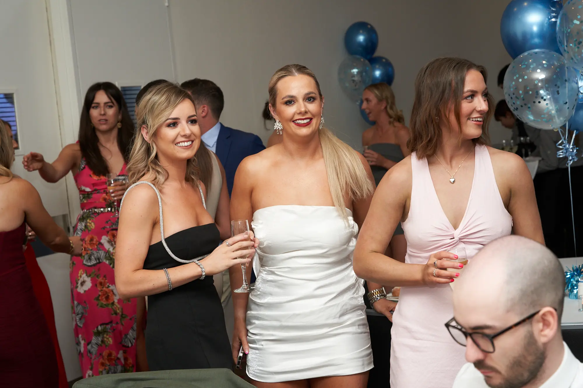 st kevins awards night pullman melbourne albert park guests socializing celebration Three women share a lively moment during St. Kevin's Awards Night at Pullman Melbourne Albert Park. The woman on the left wears a black dress with pearls and holds a champagne flute, the woman in the center wears a white strapless dress and smiles brightly at the camera, and the woman on the right wears a light pink gown, looking to her side with a joyful expression. Other guests mingle in the background, surrounded by blue and white confetti balloons. The scene captures the elegant, social, and festive energy of the evening’s celebration.