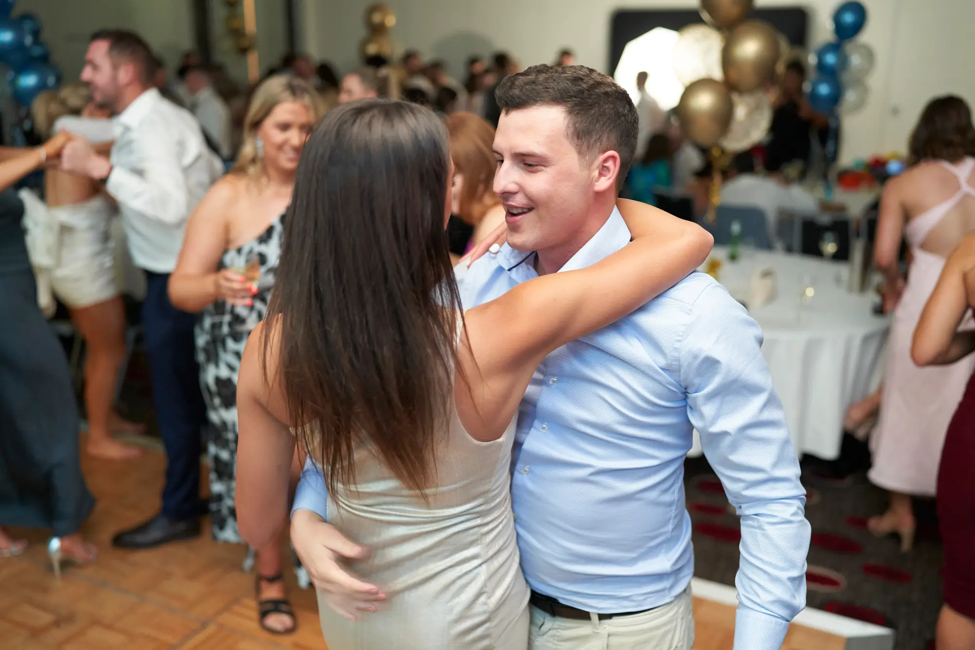 st kevins awards night pullman melbourne albert park intimate dance embrace This wide shot captures an intimate, joyful moment on the dance floor at the St. Kevin's Awards Night. A man in a light blue shirt and khaki trousers embraces a woman in a champagne satin slip dress, smiling warmly as they share a connected moment. Around them, guests in formal attire dance and mingle among tables decorated with white tablecloths and clusters of gold and blue balloons. The wooden parquet dance floor and lively atmosphere highlight the celebratory energy of the post-awards party while emphasizing the tender interaction of the central couple.