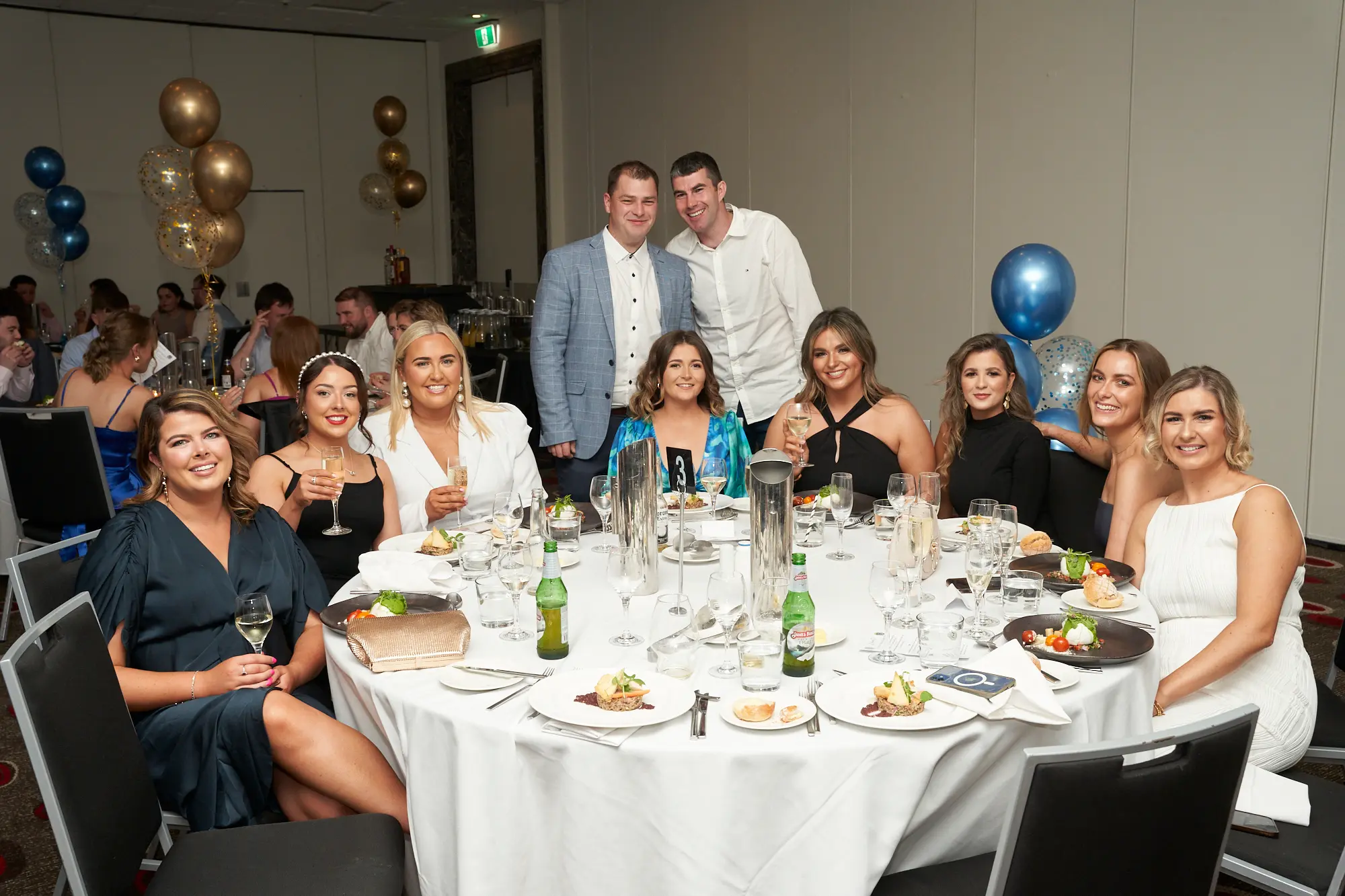 st kevins awards night pullman melbourne albert park large group dinner table Seven women seated around a dinner table, joined by two men standing behind them, smile for the camera during St. Kevin’s Awards Night at Pullman Melbourne Albert Park. The women wear formal dresses in green, black, and white, many holding wine or beer, while the men wear a grey blazer and a white shirt. The banquet hall is well-lit, with other guests at round tables in the background, decorated with blue and gold balloon clusters. The image captures the joyful, social, and celebratory atmosphere of the evening.