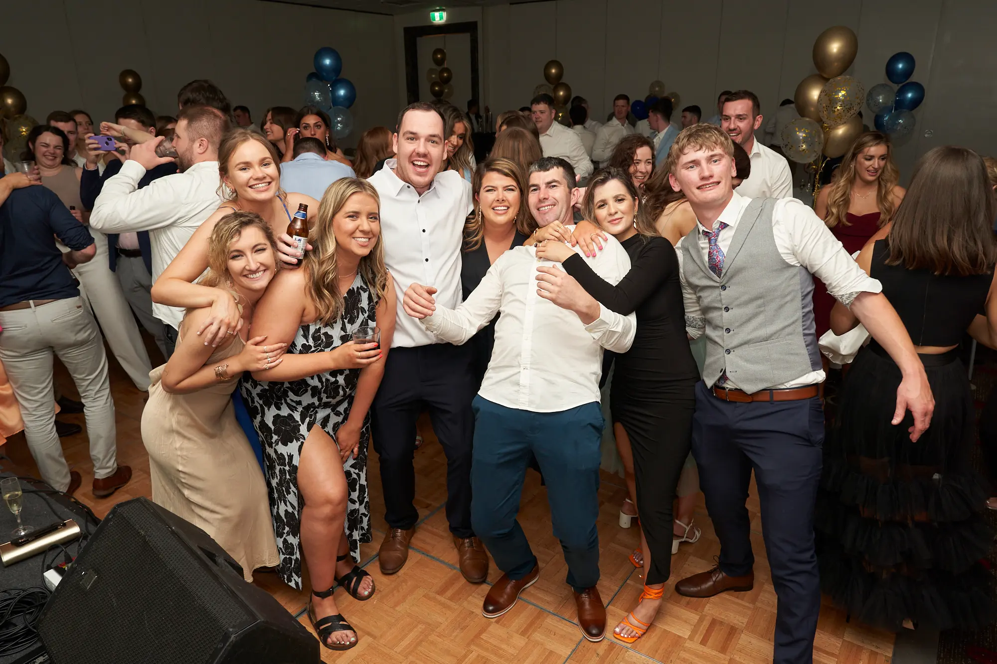 st kevins awards night pullman melbourne albert park large group friends celebrating on dancefloor A vibrant and joyful wide shot of a large group of friends posing together on the dance floor during St. Kevin’s Awards Night at Pullman Melbourne Albert Park. Around fifteen guests, dressed in elegant evening wear, smile, laugh, and raise drinks toward the camera in a spontaneous moment of celebration. The wooden dance floor glows under warm light, while gold, blue, and confetti-filled balloons decorate the background. Dinner tables and other guests mingling add depth to the festive atmosphere, capturing the pure excitement and camaraderie of the night’s celebrations.