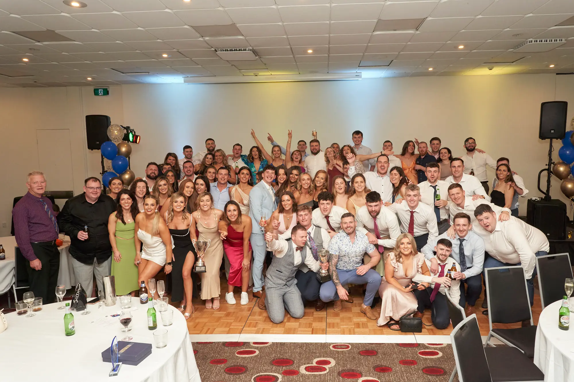st kevins awards night pullman melbourne albert park large group photo Around 50 attendees gather on the dance floor at the St. Kevin's Awards Night, dressed in formal evening wear and smiling broadly at the camera. Guests in the front row kneel or crouch, while others stand behind, some raising their hands in excitement. Two women hold clear glass awards, and a few men hold beer bottles. The wooden dance floor, dark patterned carpet, and festive clusters of gold, blue, and confetti-filled balloons frame the lively, social, and celebratory atmosphere, capturing a dynamic moment of collective joy and camaraderie during the awards night event.