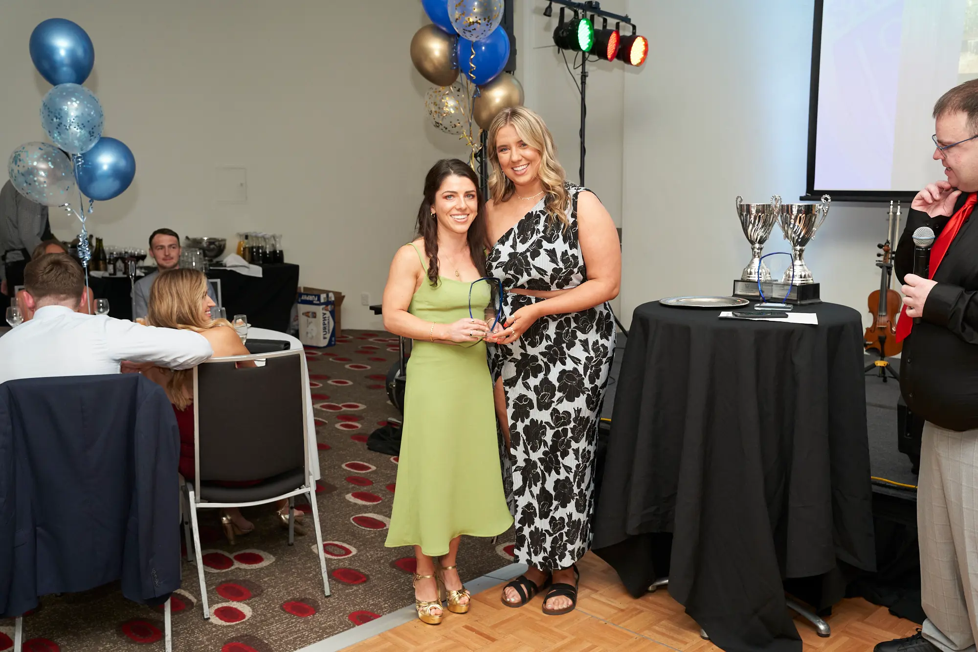 st kevins awards night pullman melbourne albert park lime green award stage Two women pose on stage at St. Kevin's GAC 2023 Awards Night at Pullman Melbourne Albert Park. The woman on the left wears a lime-green dress with gold shoes, holding a clear glass award plaque and smiling brightly. Beside her, a woman in a black-and-white floral one-shoulder dress also smiles. The stage features balloons, silver trophies, a violin, and a partially visible projection screen, creating a celebratory and formal atmosphere.
