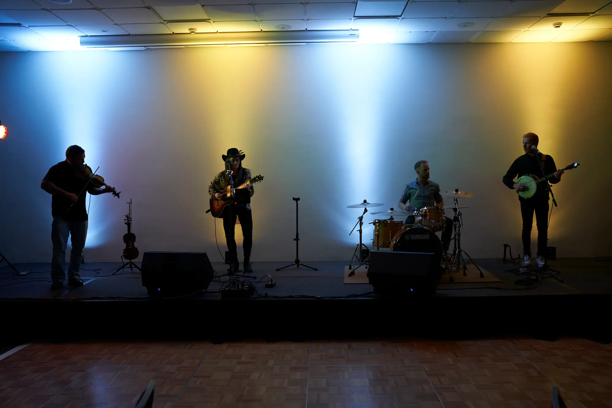 st kevins awards night pullman melbourne albert park live band performance On stage at St. Kevin's Awards Night, a lively four-piece band performs for the audience. A violinist in black plays on the left, beside a guitarist wearing a black cowboy hat. A drummer with a checkered shirt drives the rhythm behind a Yamaha drum set, while a banjo player in a black sweater adds melodic flair. Dramatic stage lighting casts cool blue and warm yellow-orange washes across the backdrop, highlighting the wooden dance floor below. The musicians’ focused expressions and energetic performance create a festive, engaging atmosphere, capturing the musical entertainment and celebratory energy of the awards night event.