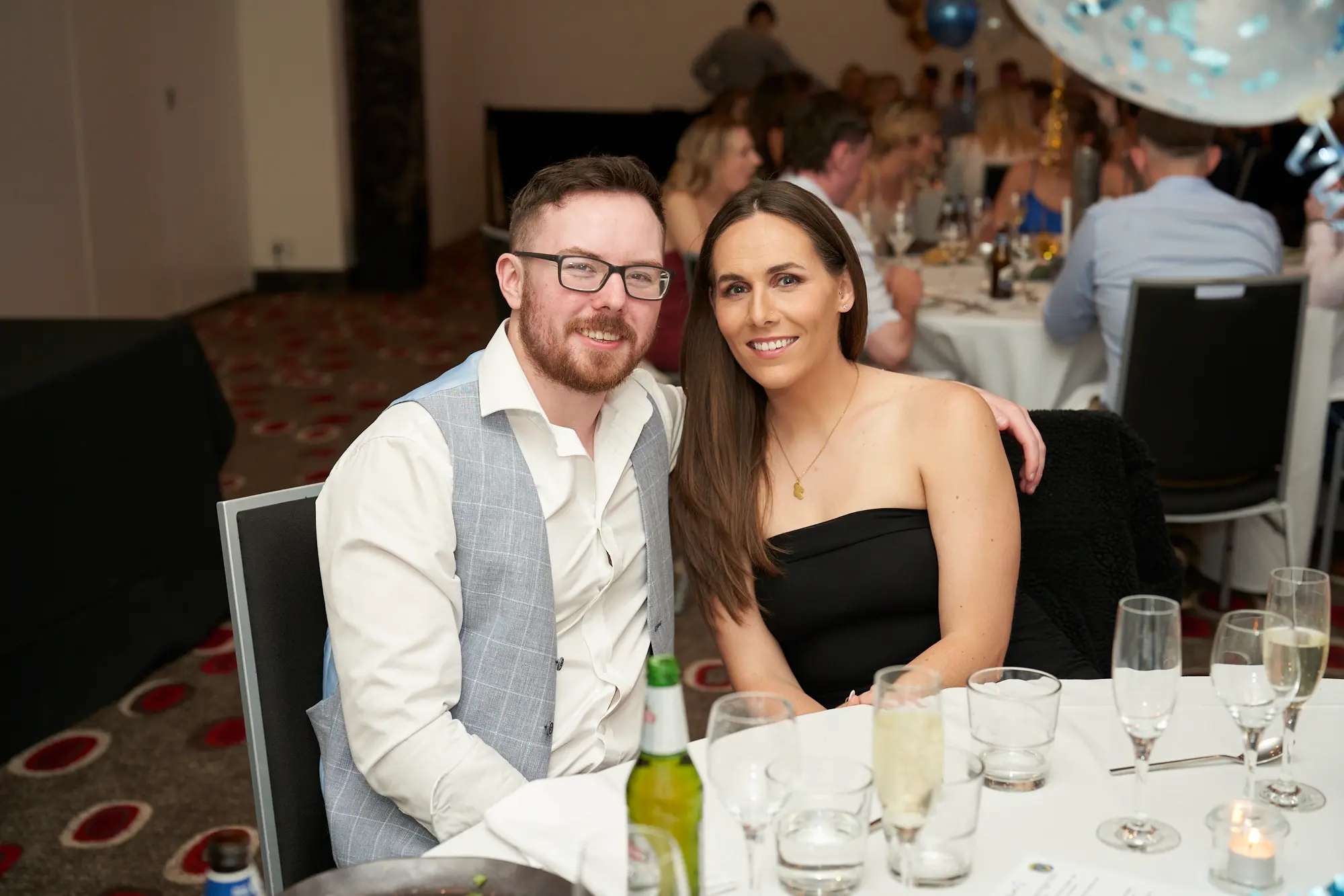 st kevins awards night pullman melbourne albert park man and woman smiling dinner table A man with a beard and glasses, wearing a light blue vest over a white shirt, smiles at the camera while seated next to a woman in a black strapless dress with a necklace during St. Kevin’s Awards Night at Pullman Melbourne Albert Park. The woman rests her left arm on the man’s shoulder, and both appear cheerful. The table is set with white linens, silverware, and glassware. In the background, other guests are seated at round tables in a banquet hall decorated with festive blue-and-white confetti balloons, capturing the joyful and celebratory atmosphere of the evening.