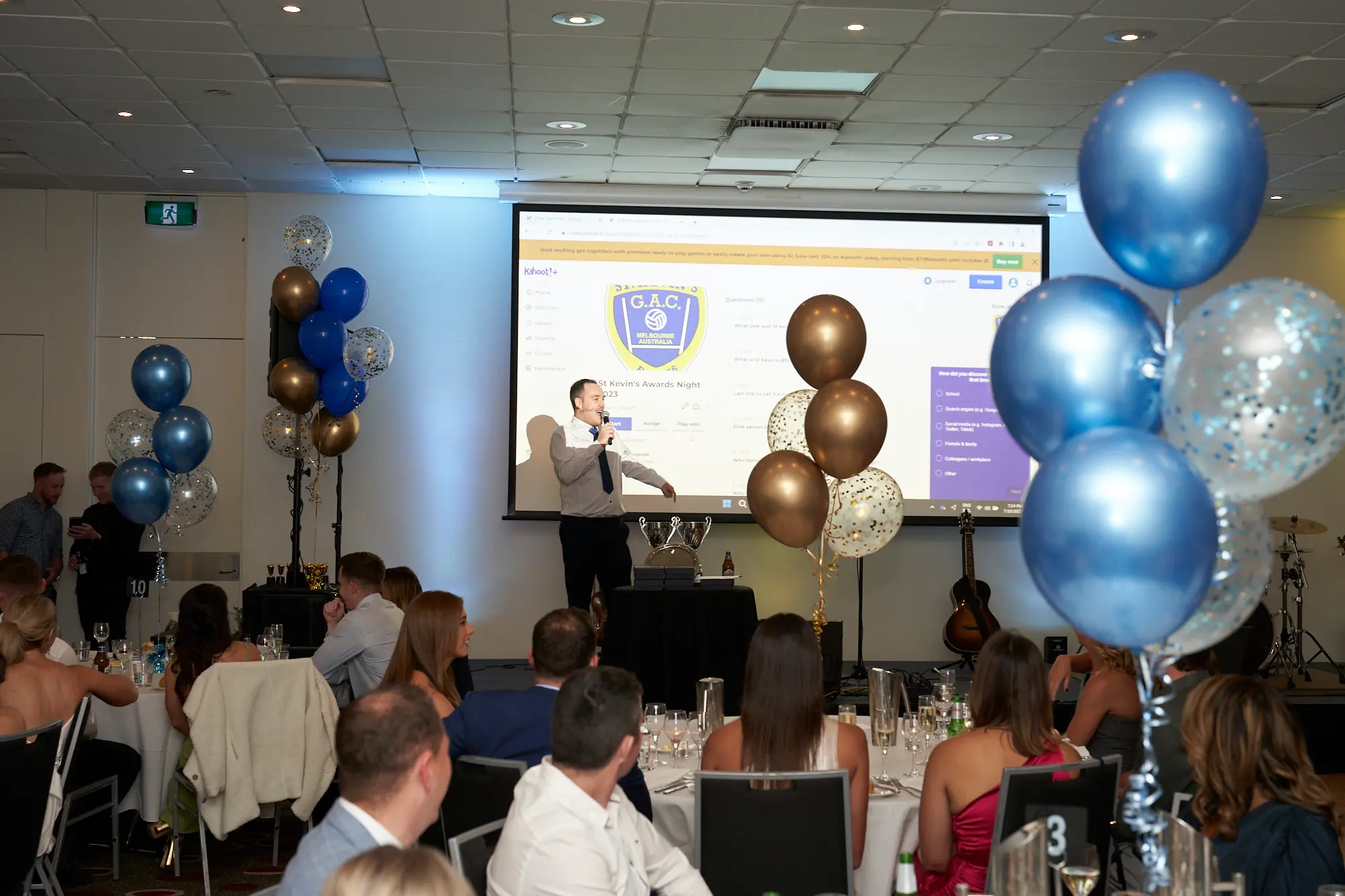 st kevins awards night pullman melbourne albert park man speaking stage A man stands on stage at St. Kevin's GAC Awards Night at Pullman Melbourne Albert Park, speaking passionately into a microphone while gesturing with his hand. The audience, dressed in formal attire, is seated at round tables covered with white tablecloths, attentively watching the presentation. Blue, gold, and confetti-filled balloons decorate the room, adding a festive touch. Behind the speaker, a large projection screen displays the "G.A.C." logo, while a guitar and partial drum set hint at live entertainment. The scene captures the formal, social, and celebratory atmosphere of a memorable awards night.