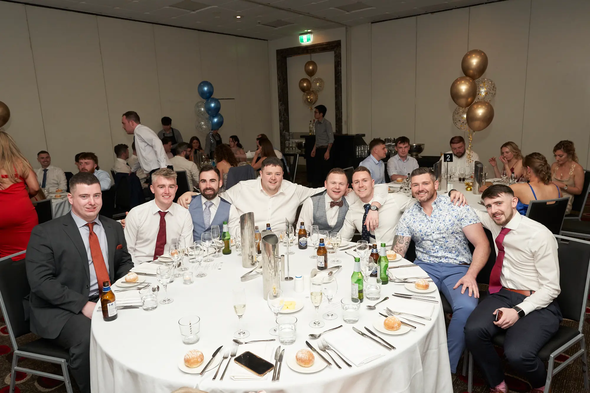 st kevins awards night pullman melbourne albert park nine men dinner table Nine men smile for the camera while seated around a dinner table during St. Kevin’s Awards Night at Pullman Melbourne Albert Park. From left to right, they wear a mix of formal attire, including suits, vests, ties, and a floral-patterned shirt, with the man on the far right holding a phone. The banquet hall in the background features other guests at round tables, dressed in formal attire, and festive blue and gold balloons, some filled with confetti. The image captures the joyful, social, and celebratory atmosphere of the awards night gathering.