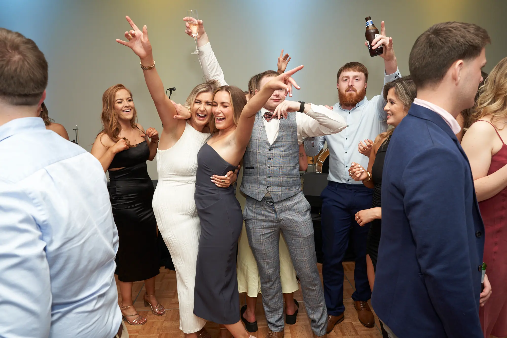 st kevins awards night pullman melbourne albert park rock on celebration This wide shot captures a spirited moment on the St. Kevin's Awards Night dance floor. A woman in white hugs a friend in dark grey while both laugh and flash "rock on" hand gestures. Nearby, a man in a grey checkered suit mirrors the gesture, and another guest raises a beer bottle in celebration. Surrounded by smiling, laughing guests holding drinks, and festive gold and blue balloons overhead, the scene radiates joyful energy, camaraderie, and the lively, celebratory atmosphere of a post-awards party where attendees are fully immersed in the fun.