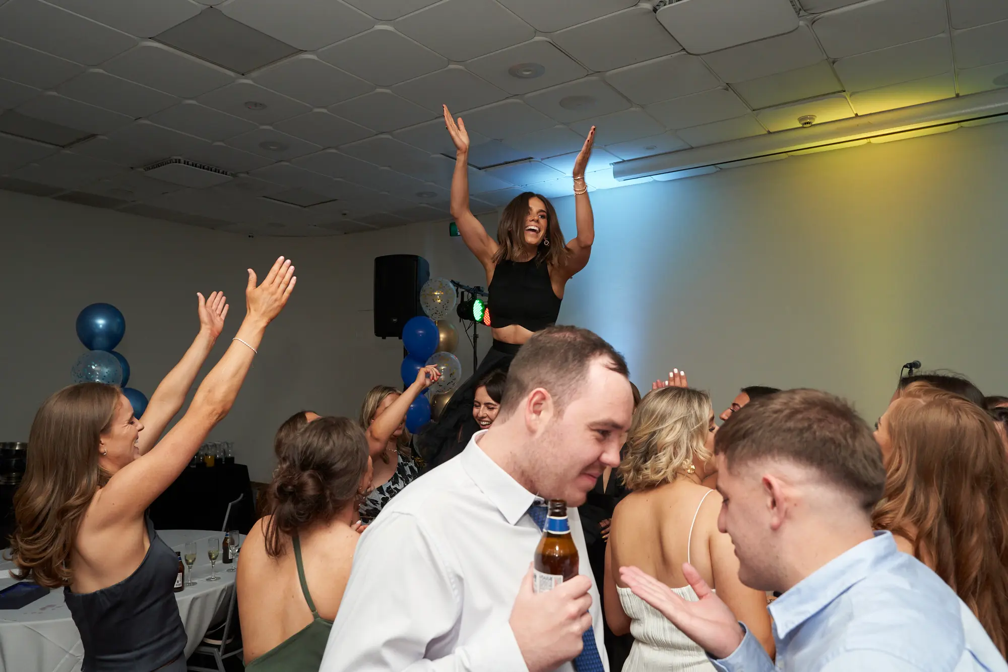 st kevins awards night pullman melbourne albert park shoulder cheer This wide shot captures a high-energy moment on the dance floor at the St. Kevin's Awards Night. A woman in a black top sits triumphantly on another guest's shoulders, laughing and raising both hands in the air. Nearby, a woman in a grey dress mirrors the excitement, while two men in the foreground engage in conversation with smiles. Festive balloons, a stage speaker, and mingling guests complete the lively scene, highlighting the celebratory, social, and joyful atmosphere of the post-awards party.