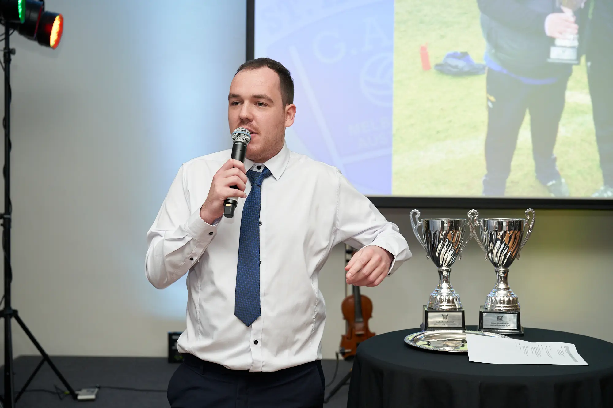 st kevins awards night pullman melbourne albert park speaker stage A man speaks into a microphone on stage at St. Kevin's GAC Awards Night at Pullman Melbourne Albert Park. He wears a white shirt and dark blue patterned tie, addressing the audience. The stage features a table with silver trophies, a violin on a stand, a projection screen displaying a trophy image, and stage lighting, creating a formal and professional awards ceremony atmosphere.