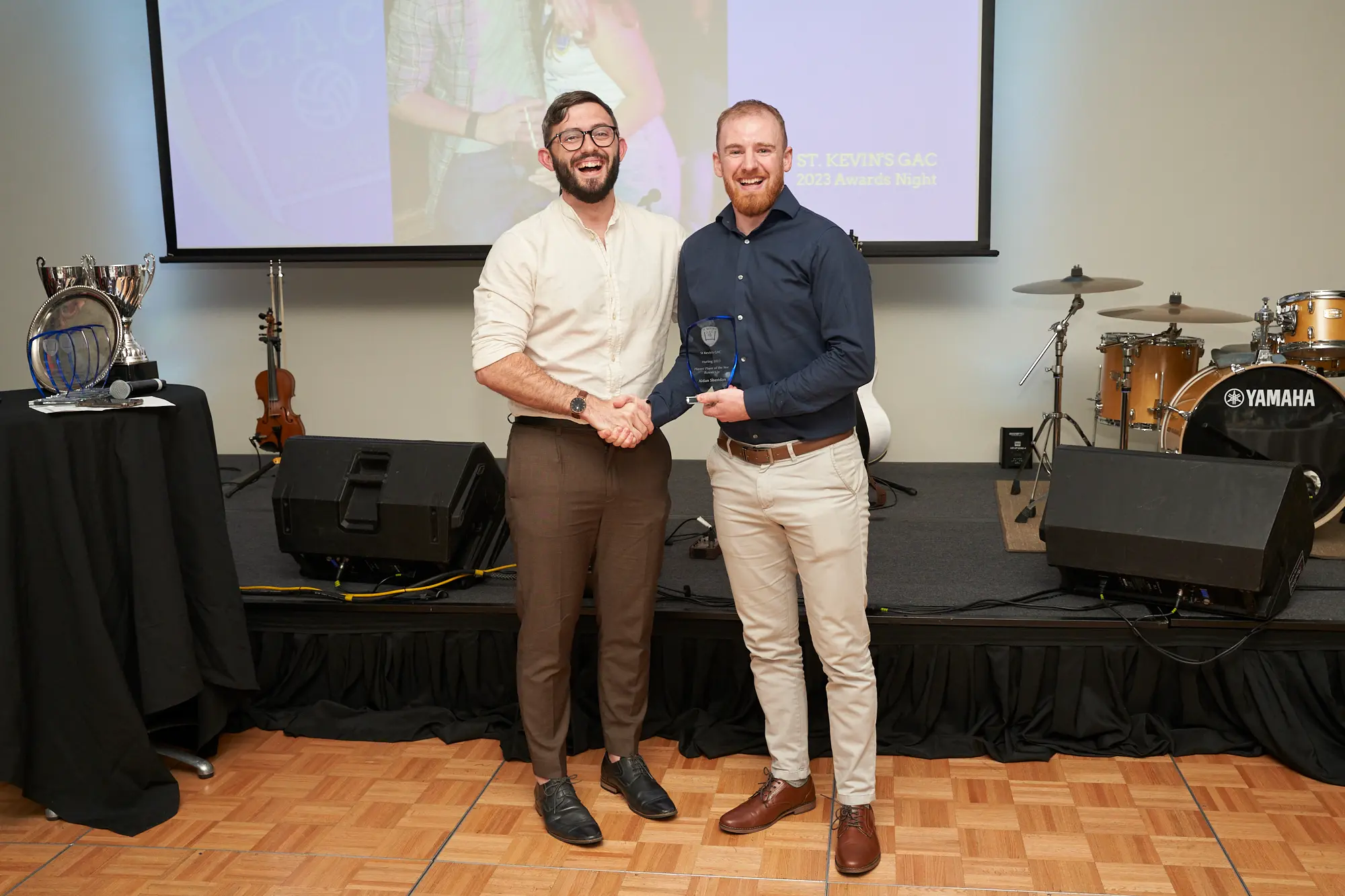 st kevins awards night pullman melbourne albert park stage award handshake navy A wide-shot image of two men on stage during St. Kevin's GAC 2023 Awards Night at Pullman Melbourne Albert Park. The man on the left wears a light-colored shirt and brown trousers, shaking hands with the winner on the right, who wears a dark navy shirt and light khaki trousers while holding a clear, shield-shaped glass award plaque. Behind them, a projection screen shows a field scene, and a Yamaha drum kit, acoustic guitar, and stage speakers are visible. Both men smile warmly, capturing a celebratory and formal moment of recognition.