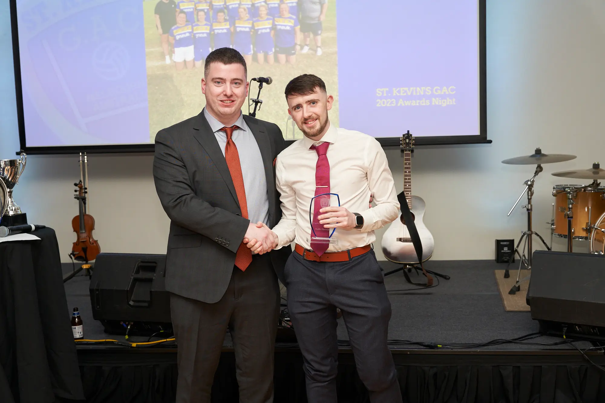 st kevins awards night pullman melbourne albert park stage award handshake A wide-shot image of two men on stage during St. Kevin's GAC 2023 Awards Night at Pullman Melbourne Albert Park. The man on the left wears a grey suit, white shirt, and rust-orange tie, shaking hands with the winner on the right, who wears a light-colored shirt, dark trousers, and a matching rust-orange tie while holding a clear glass award plaque. Behind them, a large projection screen shows a sports team, and a Yamaha drum kit, acoustic guitar, and stage speakers are visible. Both men smile warmly, capturing a celebratory and formal moment of achievement.