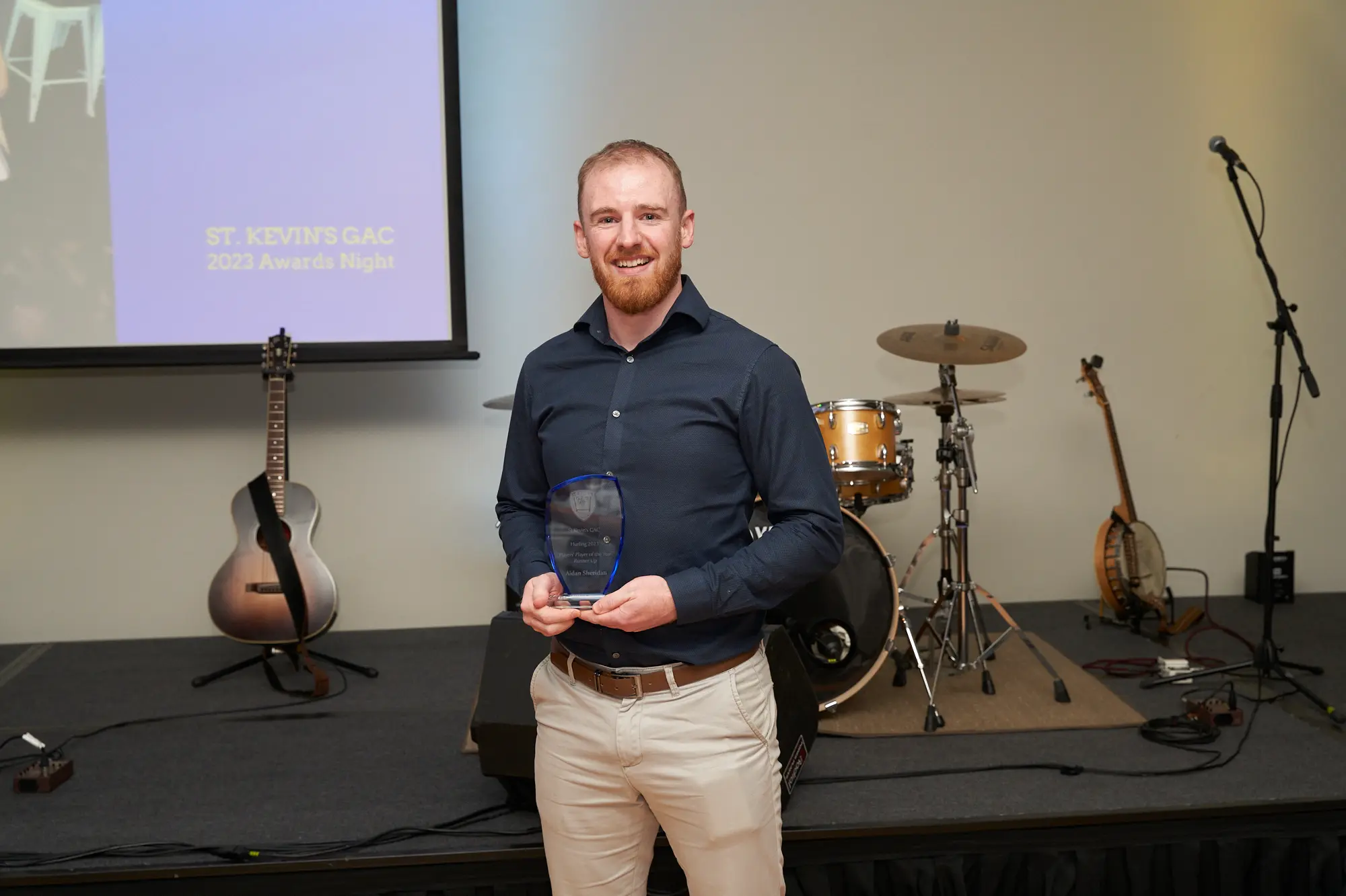 st kevins awards night pullman melbourne albert park stage award navy A wide-shot image of a man on stage at St. Kevin's GAC 2023 Awards Night at Pullman Melbourne Albert Park. He wears a dark navy button-up shirt and light khaki trousers, smiling at the camera while holding a clear, shield-shaped glass award plaque in both hands. Behind him, a projection screen confirms the event, and stage equipment including a gold drum set, cymbals, an acoustic guitar, microphones, and speakers are visible. The celebratory and formal atmosphere highlights the pride and achievement of the award recipient.