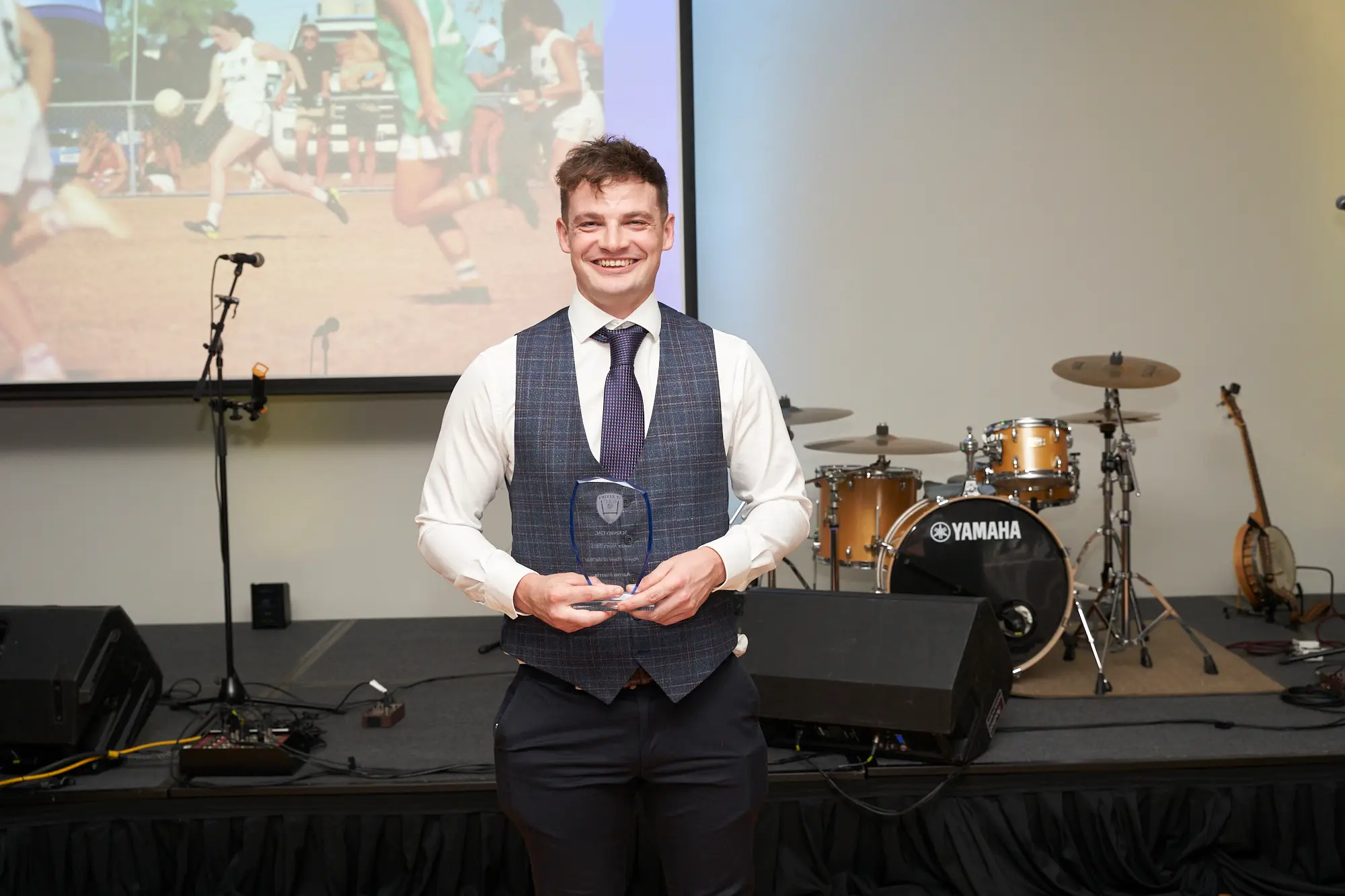 st kevins awards night pullman melbourne albert park stage award winner proud A wide-shot image of a young man standing on stage during St. Kevin's Awards Night at Pullman Melbourne Albert Park. He wears a grey checkered three-piece suit with a white shirt and dark tie, holding a clear, shield-shaped glass award plaque with both hands. Behind him, a large projection screen displays a sports team image, while a Yamaha drum set, acoustic guitar, speakers, and microphone stands are visible on the stage. The man smiles proudly, capturing a celebratory and formal moment during the awards ceremony.