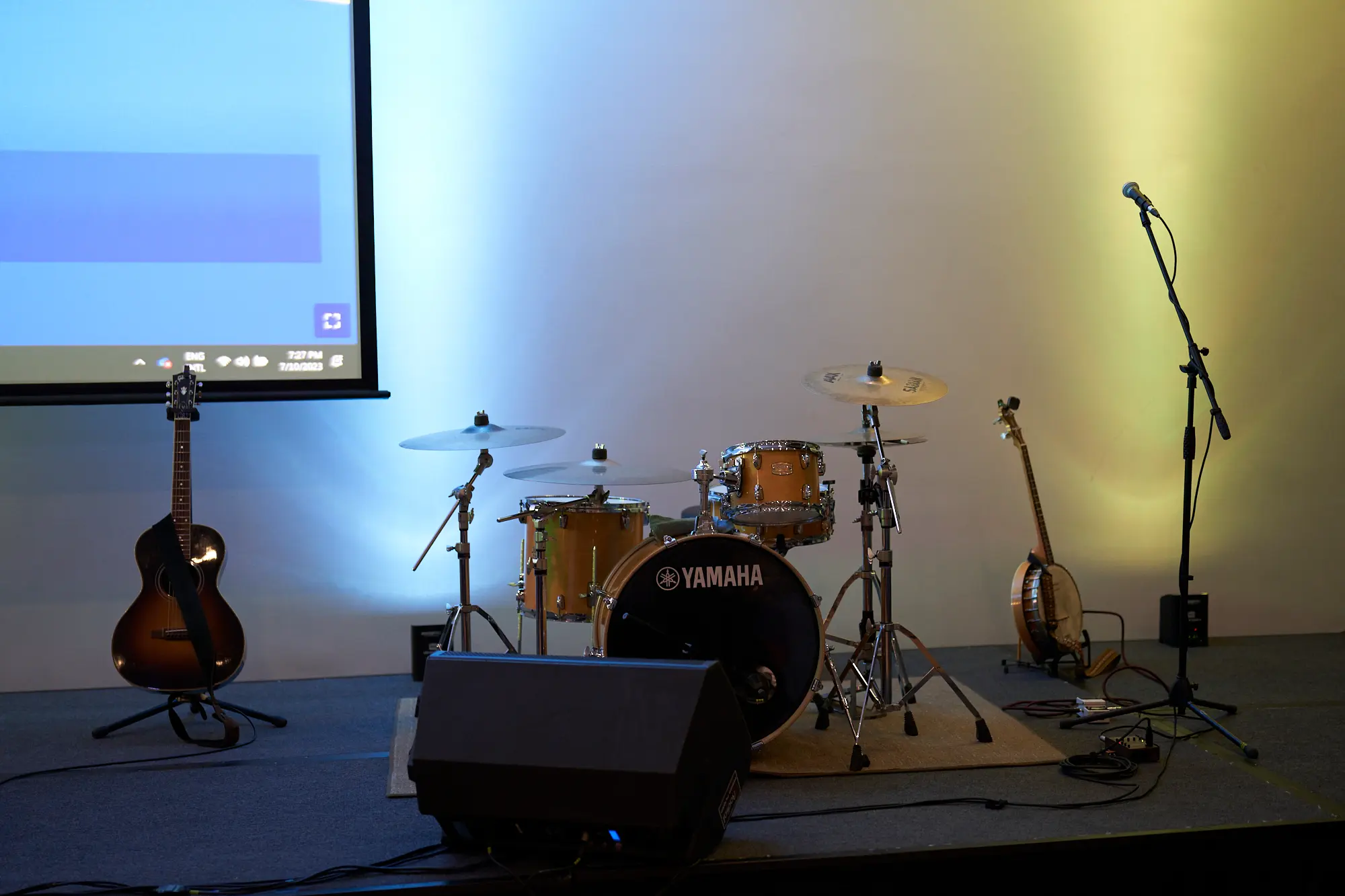 st kevins awards night pullman melbourne albert park stage setup musical instruments A stage setup at St. Kevin's Awards Night at Pullman Melbourne Albert Park features a Yamaha drum kit with a natural wood finish at its center. An acoustic guitar and a banjo rest on stands beside the drums, while a black monitor speaker sits in the foreground. The stage is illuminated by warm yellow and cool blue spotlights that highlight a white backdrop. A projector screen on the left displays a desktop interface, and a microphone on a stand waits on the right, creating an inviting scene ready for live music and performances during the evening celebration.