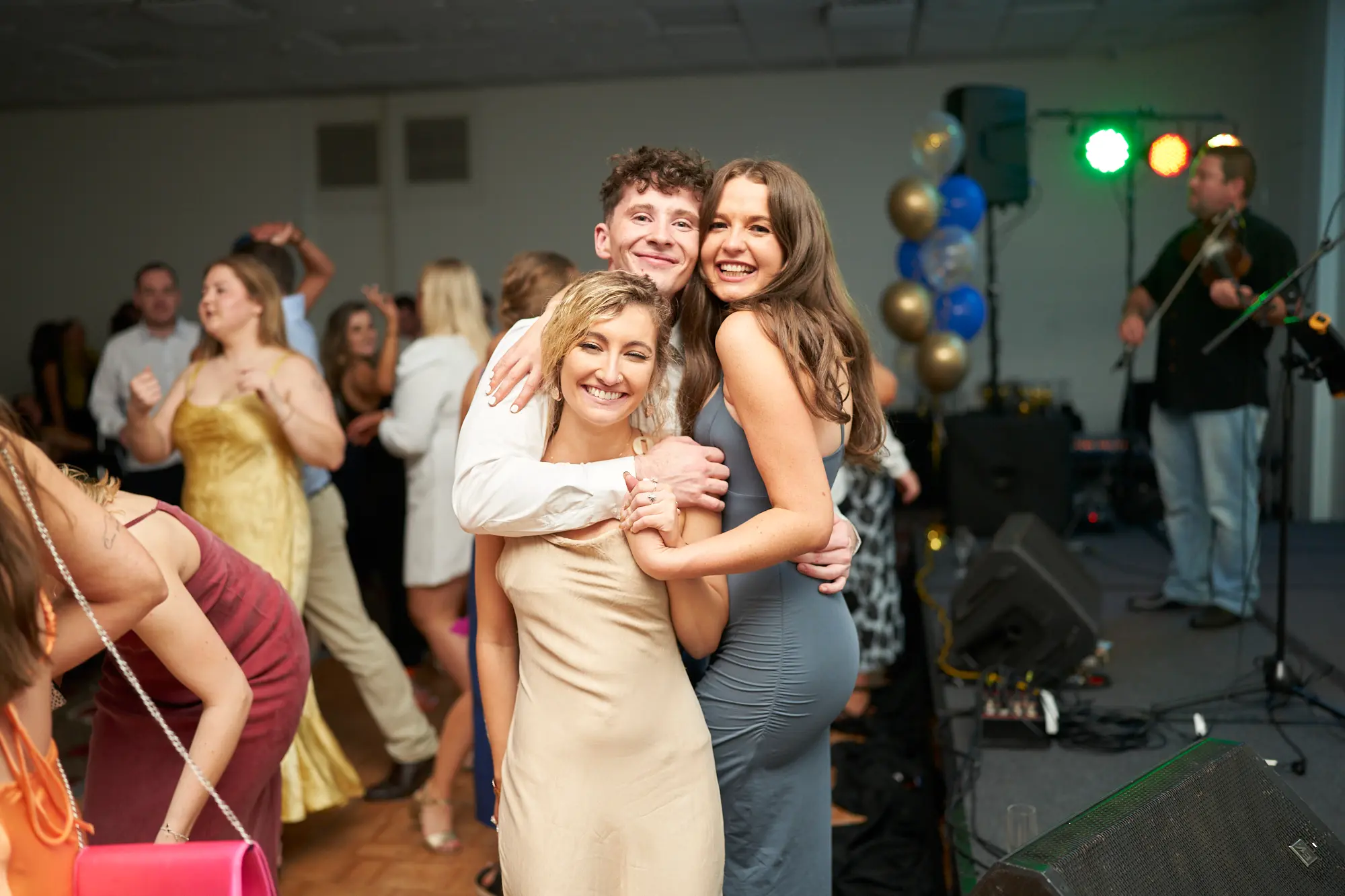 st kevins awards night pullman melbourne albert park three friends dancing celebration Three young adults pose joyfully on the dance floor during St. Kevin’s Awards Night at Pullman Melbourne Albert Park. The man in the center, wearing a white shirt, wraps his arms around two women — one in a light beige satin dress and the other in a grey bodycon slip dress — as they smile brightly at the camera. Behind them, guests mingle and dance under warm light, while a live band plays on a nearby stage decorated with gold and blue balloons. The lively atmosphere captures the warmth, fun, and togetherness of the evening’s celebration.