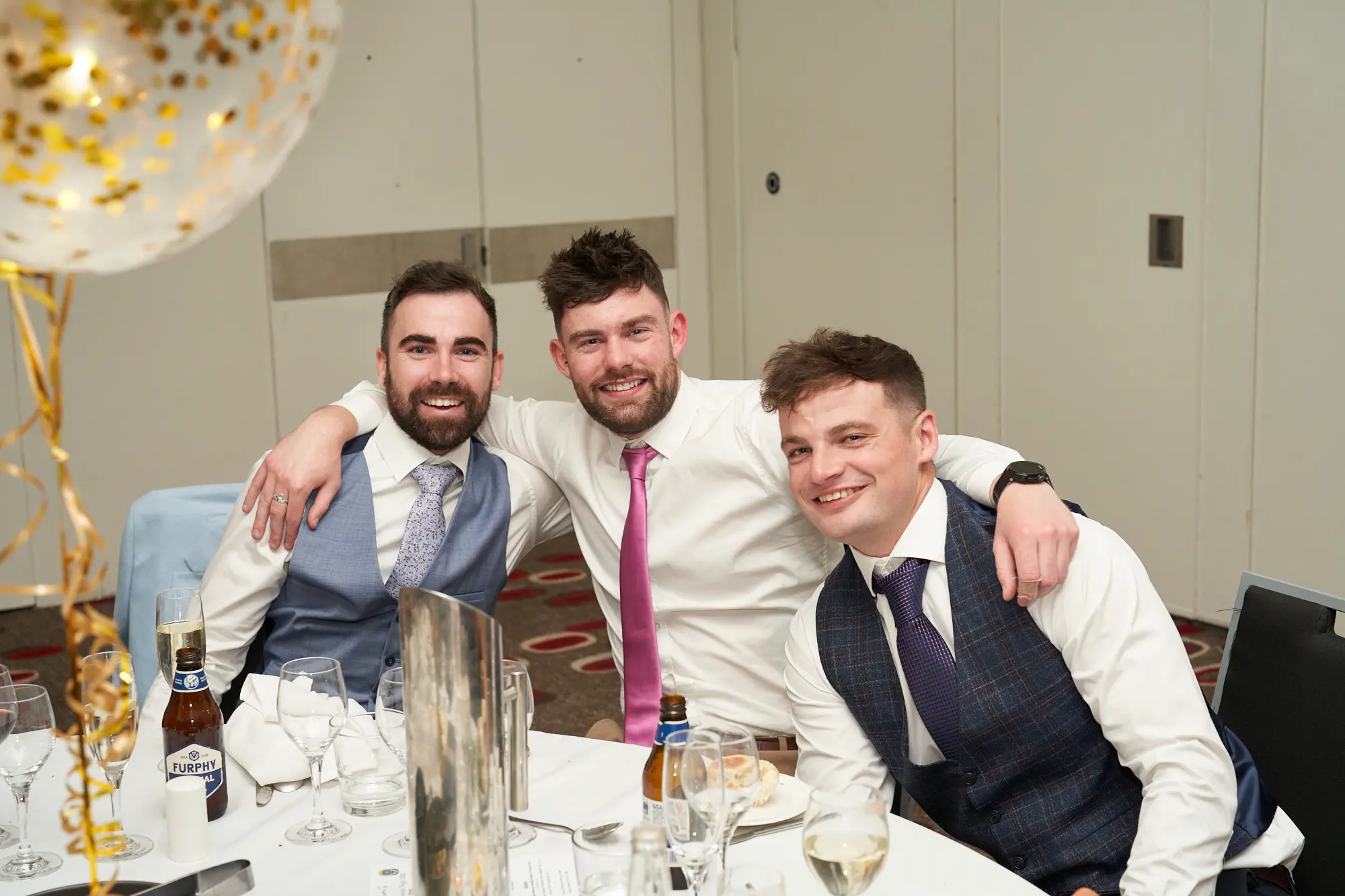 st kevins awards night pullman melbourne albert park three men smiling dinner table Three men smile for the camera while seated together at a dinner table during St. Kevin’s Awards Night at Pullman Melbourne Albert Park. The man in the center wears a white shirt and pink tie, with his arms around the shoulders of the men on either side, who wear blue patterned vests and ties. A bottle of Furphy beer sits on the table in front of them. Behind them, the banquet hall features light-colored walls, white tablecloths, and a large transparent balloon filled with gold confetti, capturing the joyful and festive atmosphere of the evening celebration.