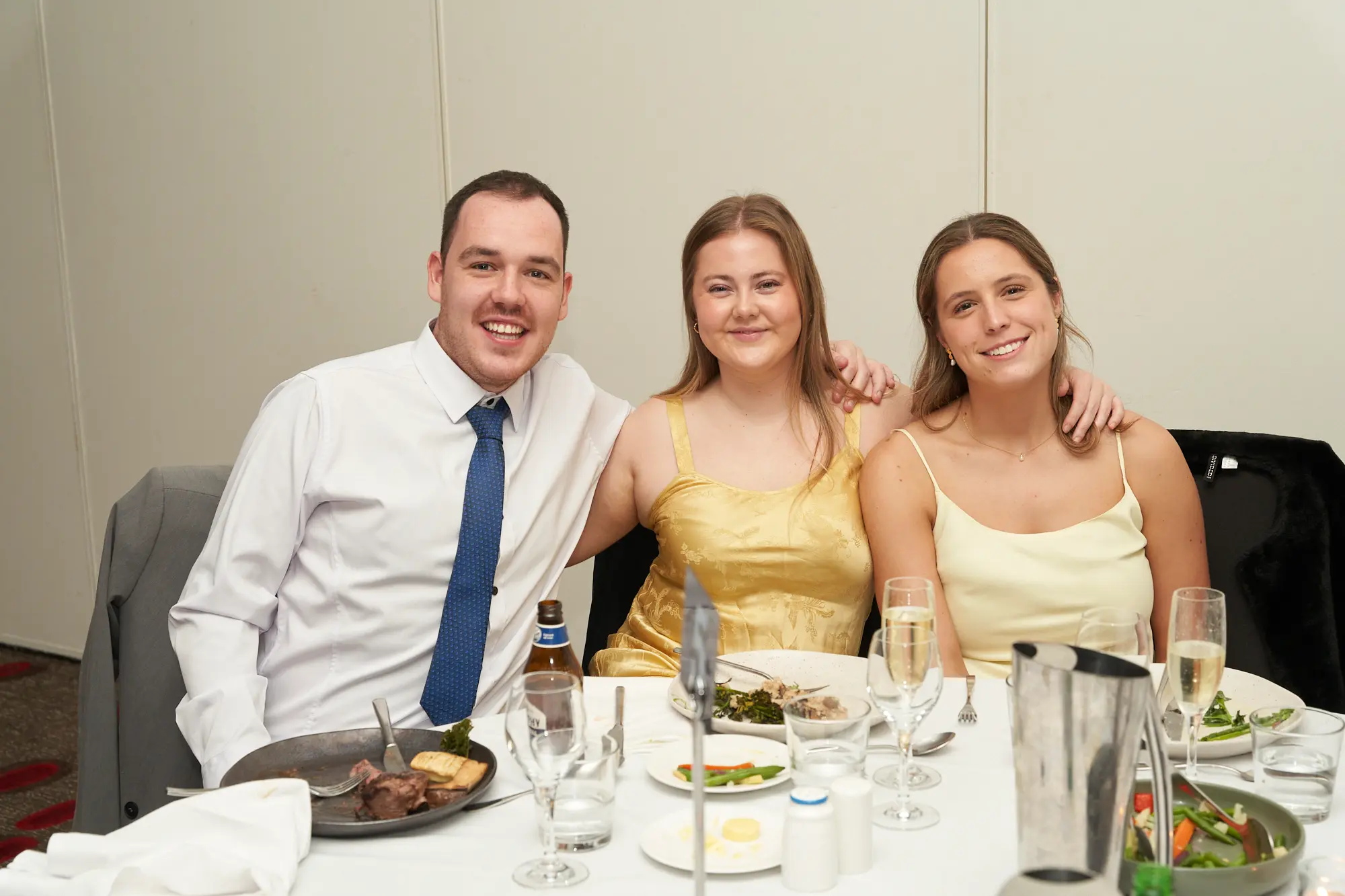 st kevins awards night pullman melbourne albert park three people smiling dinner table Three people smile warmly for the camera while seated together at a dinner table during St. Kevin’s Awards Night at Pullman Melbourne Albert Park. The man on the left wears a white shirt and blue tie with his arm around the woman in the center, who wears a gold dress, while the woman on the right wears a light yellow spaghetti-strap gown. The table is set with plates, glasses, and drinks, and the banquet hall features light-colored walls and a red patterned carpet. The image captures the friendly, joyful, and celebratory atmosphere of the evening.