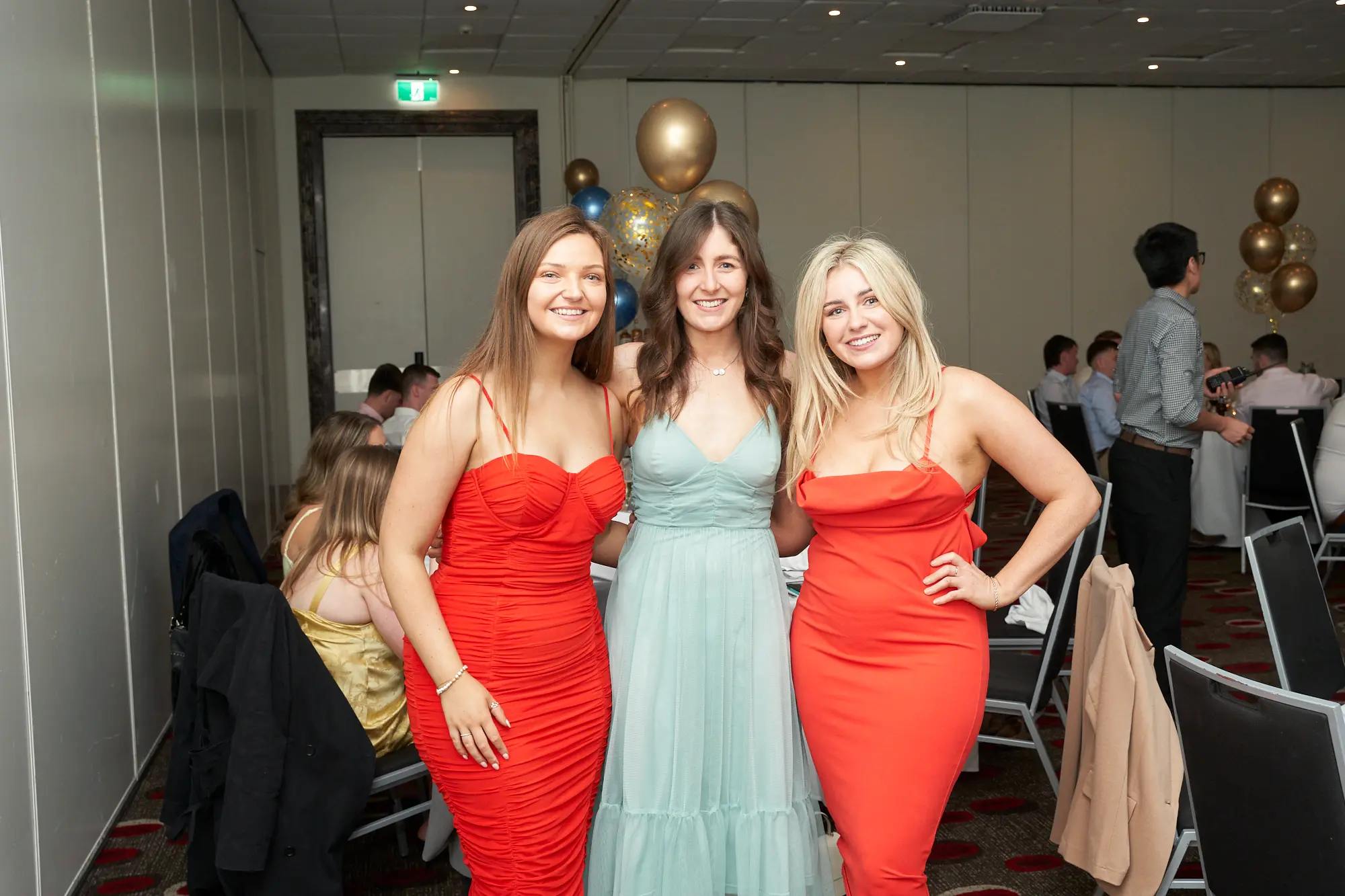 st kevins awards night pullman melbourne albert park three women posing banquet hall Three women smile for a photo at St. Kevin's Awards Night at Pullman Melbourne Albert Park. The woman on the left wears a red strapless dress, the center woman wears a mint green V-neck gown with a tiered skirt, and the woman on the right wears a red spaghetti-strap dress with a cowl neck, her hand on her hip. Behind them, guests sit at round tables with white linens and black chairs, surrounded by gold and blue balloon decorations. The scene radiates elegance, joy, and the celebratory spirit of the awards night gathering.