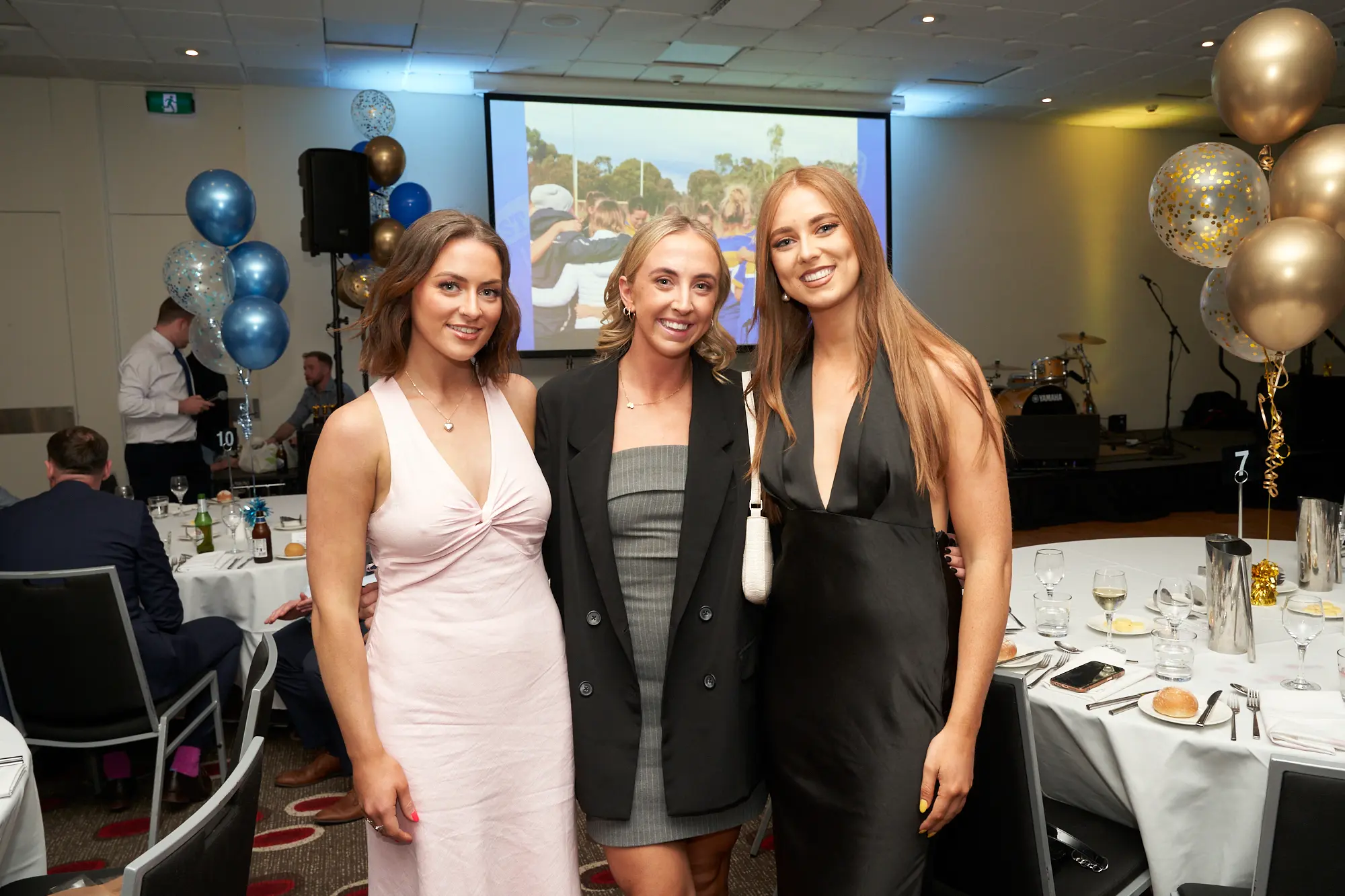 st kevins awards night pullman melbourne albert park three women posing celebration Three women stand side by side, smiling for the camera at St. Kevin's Awards Night at Pullman Melbourne Albert Park. The woman on the left wears a light pink sleeveless dress, the center woman wears a strapless grey dress paired with a black blazer, and the woman on the right wears a black V-neck gown. Behind them, a projector screen displays an image of a team celebrating, while blue and gold balloons frame the stage with musical instruments and speakers. The scene conveys a joyful, elegant, and festive atmosphere that highlights the evening’s sense of celebration and camaraderie.