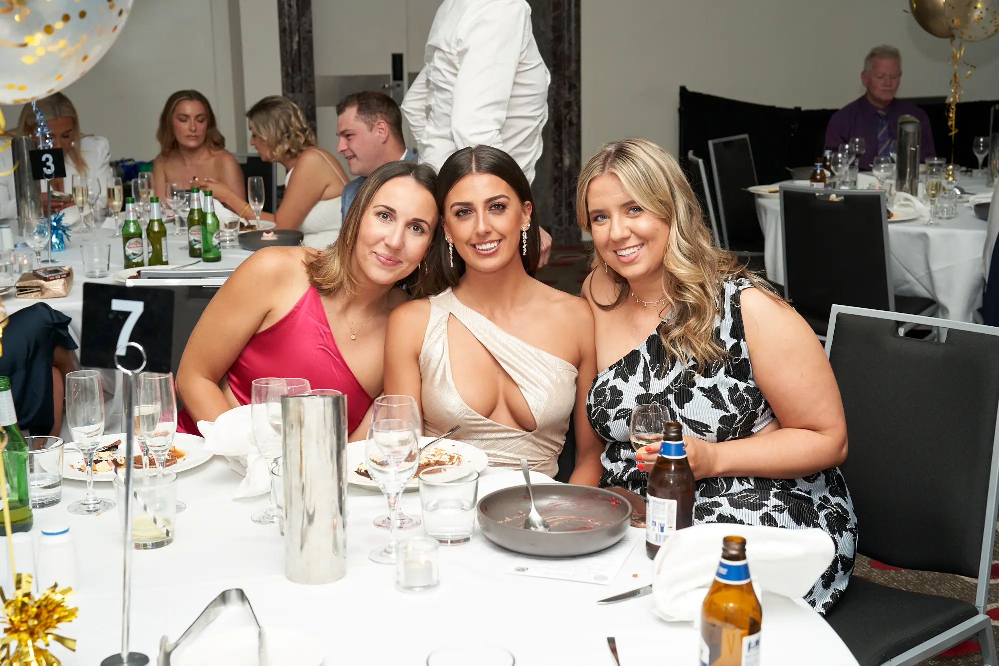 st kevins awards night pullman melbourne albert park three women seated dinner table Three women smile warmly for the camera while seated together at a dinner table during St. Kevin's Awards Night at Pullman Melbourne Albert Park. The woman on the left wears a pink satin dress, the center woman wears a champagne-colored one-shoulder gown with a low neckline, and the woman on the right wears a black and white patterned one-shoulder dress, holding a glass of wine and a bottle of beer. Behind them, guests dine and chat in a lively banquet hall decorated with balloons and white table settings, capturing the festive and joyful atmosphere of the evening celebration.