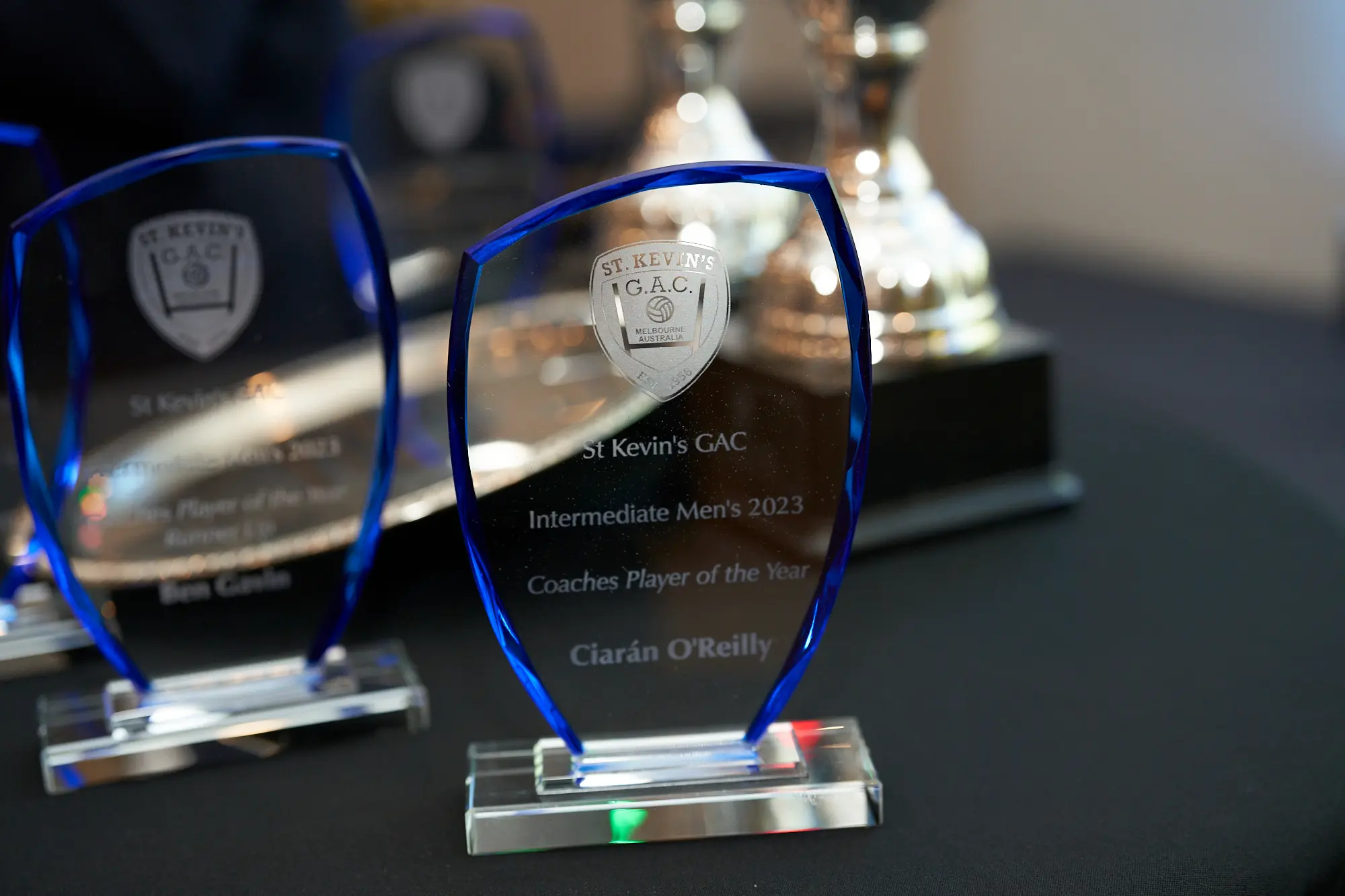 st kevins awards night pullman melbourne albert park trophies display coaches player of the year A close-up of two elegant trophies displayed at St. Kevin's Awards Night at Pullman Melbourne Albert Park. The foreground trophy, made of clear glass with blue-tinted edges and a silver base, features the St. Kevin’s G.A.C. logo and is engraved with “Intermediate Men’s 2023 – Coaches Player of the Year – Ciarán O’Reilly.” Behind it, another similar trophy inscribed with “Ben Goslin” is slightly out of focus. Several more awards glimmer in the background, creating a proud and polished display that celebrates sporting excellence and achievement.