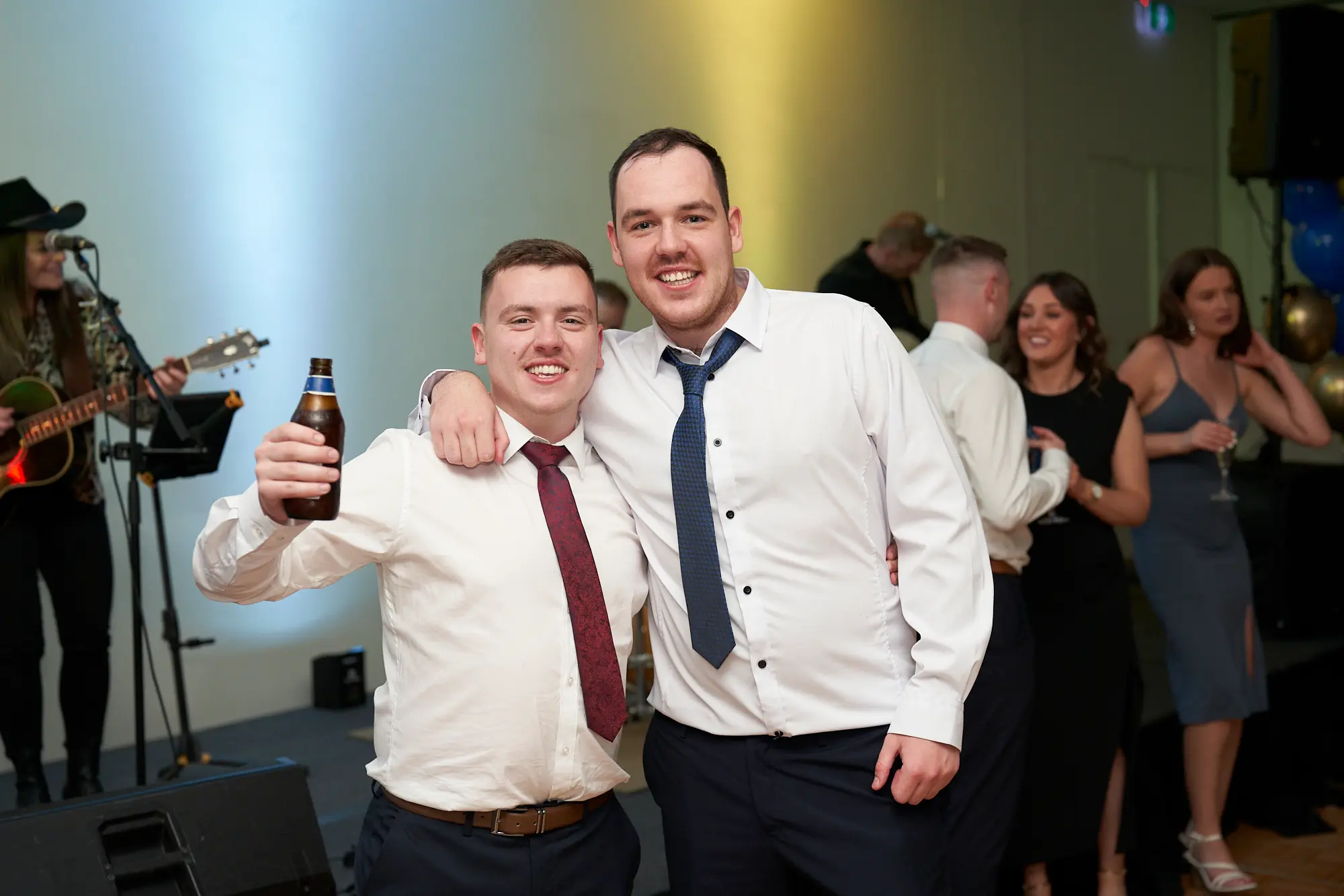 st kevins awards night pullman melbourne albert park two friends live band celebration Two young men smile warmly for the camera during St. Kevin’s Awards Night at Pullman Melbourne Albert Park. Both wear crisp white shirts and ties, standing close together with their arms around each other in a friendly pose. One holds a beer bottle while behind them, a live band performs on stage — a woman in a cowboy hat plays an acoustic guitar amid microphones and speakers. Other formally dressed guests mingle and chat near blue and gold balloons. The lively scene captures a joyful and relaxed celebration filled with laughter, music, and camaraderie.