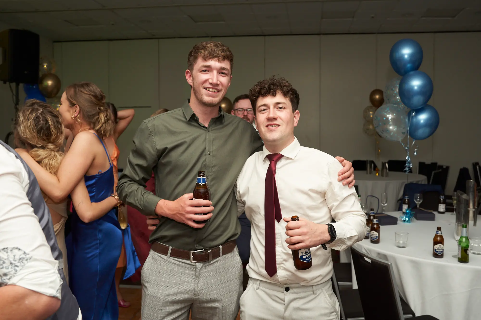 st kevins awards night pullman melbourne albert park two men celebrating with drinks Two young men share a friendly moment during St. Kevin’s Awards Night at Pullman Melbourne Albert Park. Standing side by side with arms around each other’s shoulders, they smile warmly at the camera, each holding a beer bottle. One wears an olive-green shirt and grey checkered trousers, while the other sports a white shirt, maroon tie, and white trousers. Behind them, guests mingle around tables set with drinks and glassware, surrounded by gold and blue balloons. The atmosphere is joyful, relaxed, and festive, capturing the camaraderie and fun of the evening’s celebrations.