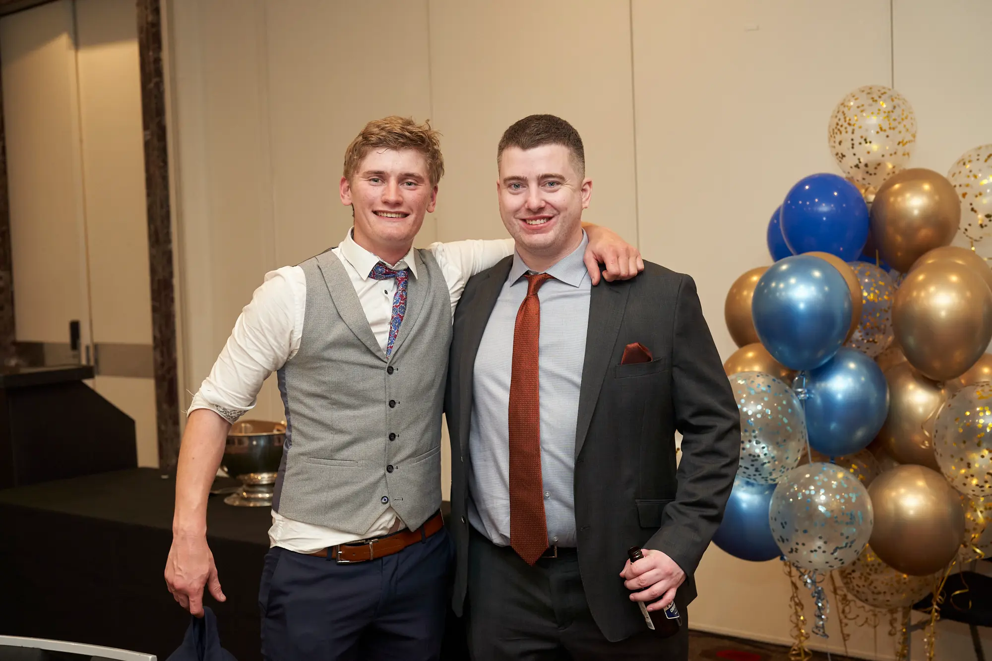 st kevins awards night pullman melbourne albert park two men celebrating with trophy and balloons A wide shot of two young men posing together during St. Kevin’s Awards Night at Pullman Melbourne Albert Park. Both are smiling brightly at the camera — one wearing a white shirt with a grey checkered waistcoat and navy trousers, the other in a dark grey suit with a rust-orange tie. They stand close, one man’s arm around the other’s shoulder, exuding warmth and camaraderie. Behind them, a large display of gold and blue balloons adds a festive backdrop, while a silver trophy glints on a nearby table, symbolizing the night’s celebratory and award-filled atmosphere.
