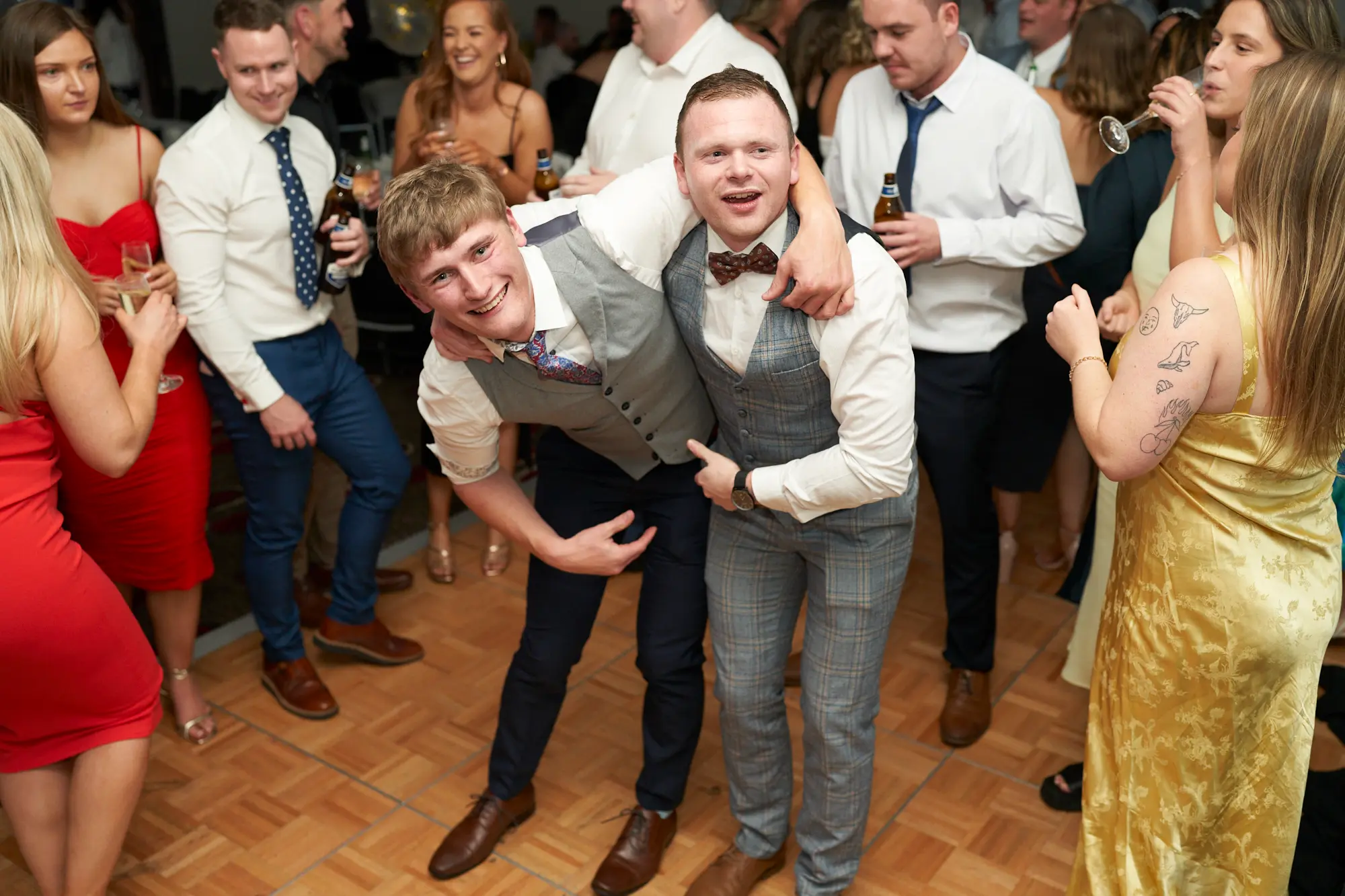 st kevins awards night pullman melbourne albert park two men dance floor celebration Two young men pose together on the wooden dance floor during St. Kevin’s Awards Night at Pullman Melbourne Albert Park. The man on the left, in a white shirt, grey waistcoat, and navy trousers, leans in with a big smile and his arm around his friend. The man on the right, wearing a grey checkered waistcoat with a patterned bowtie, points playfully toward the ground while smiling brightly. Behind them, guests mingle, chat, and enjoy drinks under warm evening lighting, with women in gold and red dresses adding to the lively, festive energy of the awards celebration.