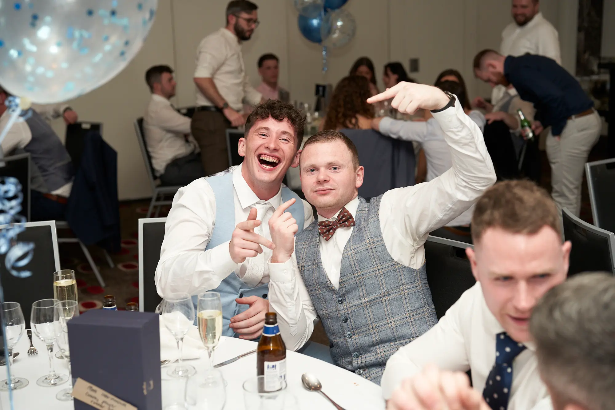 st kevins awards night pullman melbourne albert park two men laughing table Two men share a playful moment while seated at a dinner table during St. Kevin's Awards Night at Pullman Melbourne Albert Park. The man on the left laughs heartily, pointing at the camera with both hands, while the man on the right grins, wearing a plaid vest and bow tie, also pointing enthusiastically. Behind them, guests mingle and dine at round tables in a bright banquet hall decorated with confetti-filled balloons. The scene captures the warmth, humor, and lively spirit of an evening filled with laughter and celebration among friends.