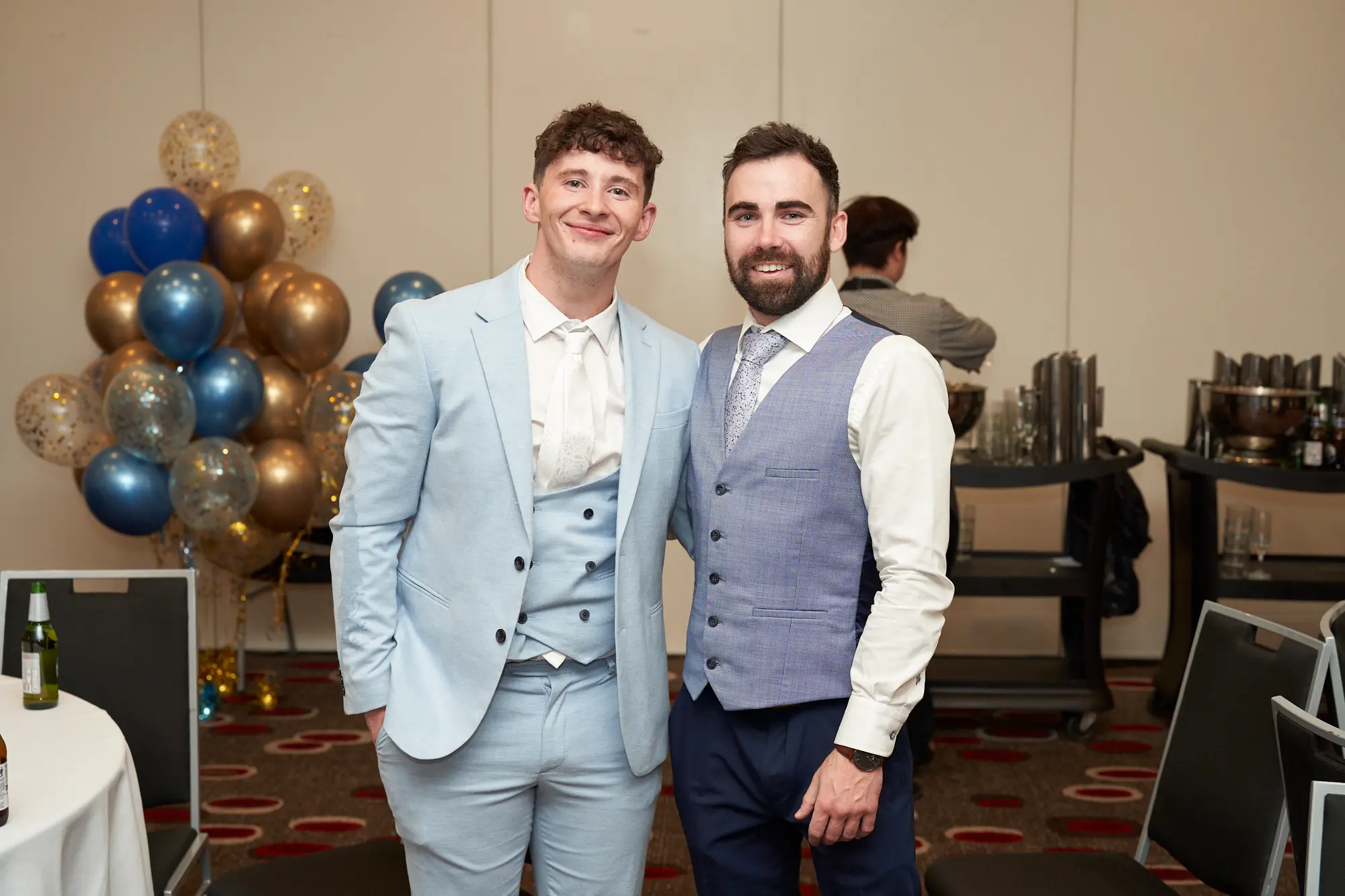 st kevins awards night pullman melbourne albert park two men posing together celebration A wide shot of two men posing together during St. Kevin’s Awards Night at Pullman Melbourne Albert Park. Both are dressed in elegant formal wear — one in a light blue three-piece suit with a matching tie, the other in a white shirt with a patterned blue-grey waistcoat and navy trousers. They stand close with friendly smiles, one man’s arm around the other’s shoulder, showing genuine camaraderie. Behind them, gold and blue confetti-filled balloons add a festive touch, while tables with white linens and drinks complete the lively, celebratory atmosphere of the evening.