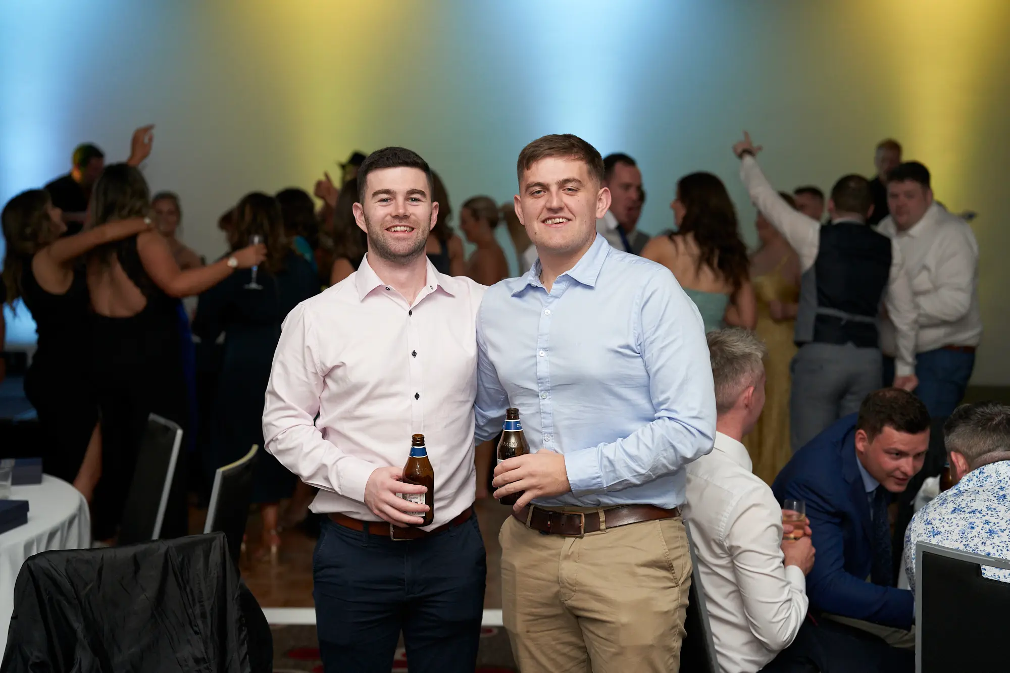 st kevins awards night pullman melbourne albert park two men social gathering Two young men pose together during the St. Kevin's Awards Night, surrounded by a lively crowd. The man on the left wears a light pink button-up shirt with dark navy trousers, holding a Corona beer, while the man on the right wears a light blue shirt and khaki trousers, also holding a beer. Behind them, guests in formal attire dance, clap, and chat under blue and yellow stage lighting. The room features a dark patterned carpet, wooden dance floor, and beige walls. The joyful, social energy reflects the celebratory spirit of the awards night.