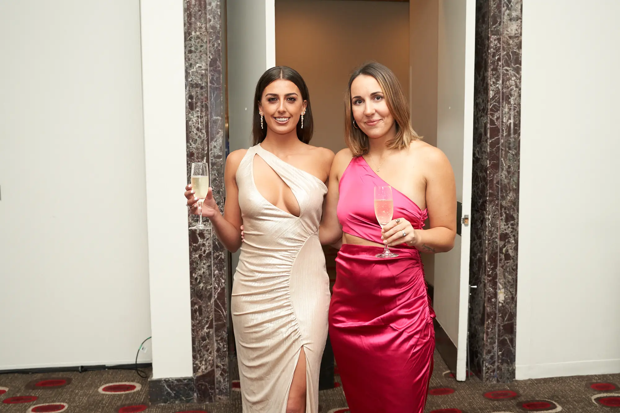 st kevins awards night pullman melbourne albert park two women guest arrival Two elegantly dressed women pose side by side at St. Kevin's Awards Night at Pullman Melbourne Albert Park, holding champagne glasses and smiling warmly at the camera. The woman on the left wears a champagne-colored, one-shoulder gown with a ruched bodice and high slit, while the woman on the right wears a magenta one-shoulder dress with a waist cutout. The marble-patterned wall and dark wooden door in the background suggest a luxurious hotel venue, creating a glamorous and celebratory atmosphere that captures the spirit of guest arrivals at this formal event.