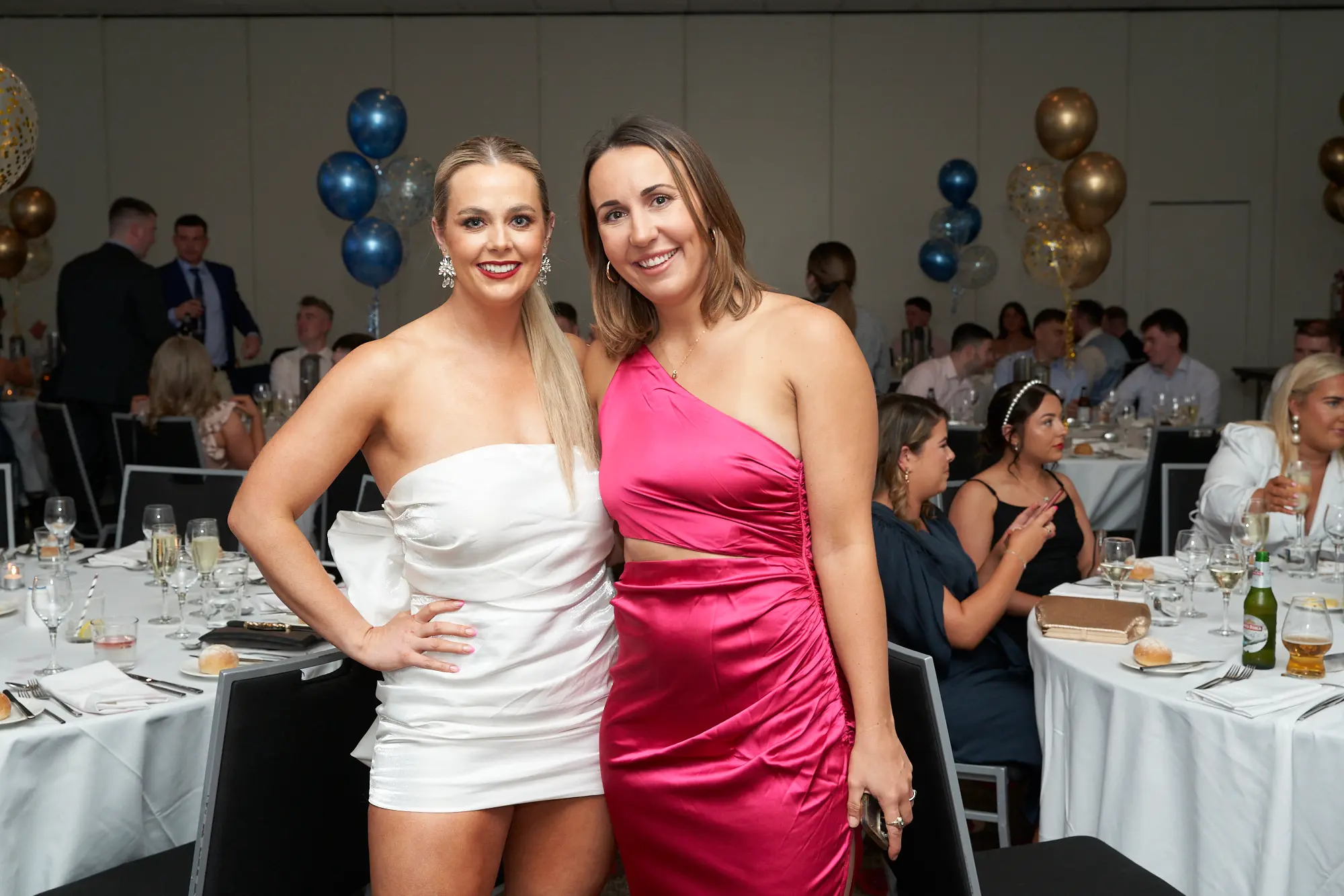 st kevins awards night pullman melbourne albert park two women posing banquet hall Two women pose cheerfully for a photo at St. Kevin's Awards Night at Pullman Melbourne Albert Park. The woman on the left wears a white strapless mini dress with a large bow at the back, her hand on her hip, while the woman on the right wears a shiny magenta one-shoulder dress with a waist cutout, her arm around her friend’s shoulder. Behind them, guests sit at round tables covered in white linens, surrounded by blue, gold, and white balloons. The warm lighting and festive décor capture the elegant and joyful atmosphere of the evening celebration.